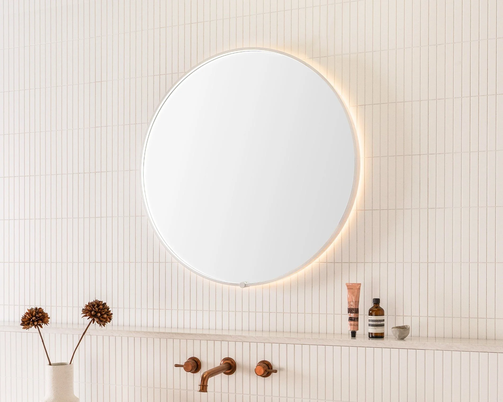 Round illuminated mirror on a white tiled bathroom wall with a beige shelf below holding a vase with dried flowers, skincare products, and a small stone.