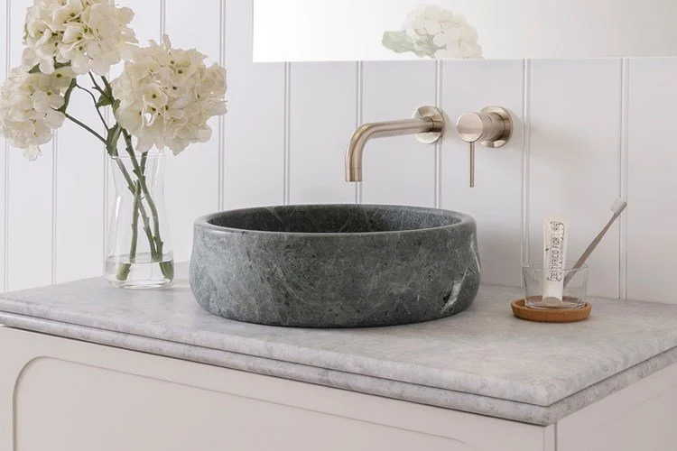 A bathroom sink area featuring a round gray stone basin, a brass wall-mounted faucet, a glass vase with white hydrangeas, and a toothbrush holder with a toothbrush on a marble countertop.