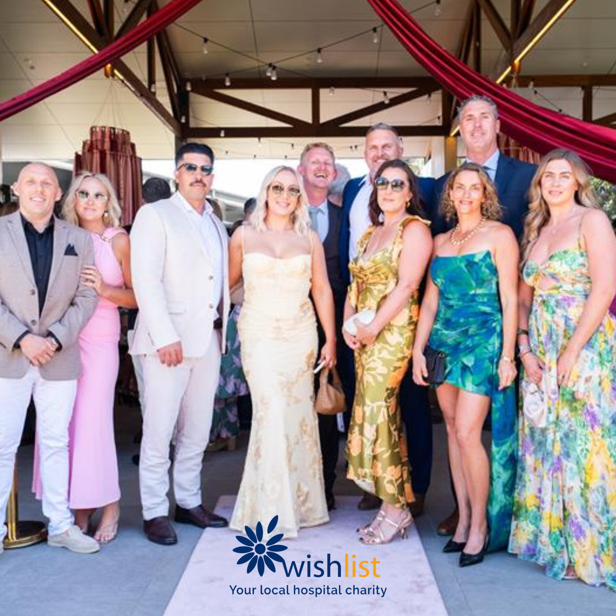 Group of people dressed in formal attire at an event organized by wish list, a local hospital charity, with colorful decorations and a wooden pavilion above.