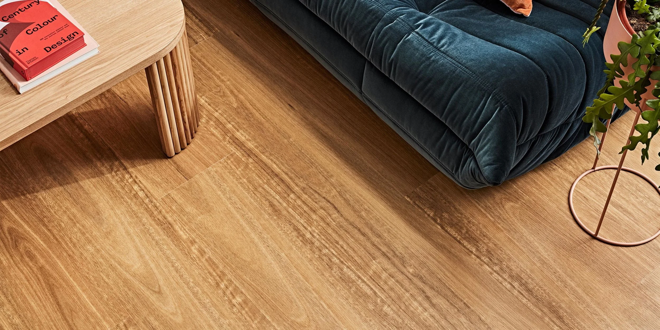 Close-up of a wooden floor, part of a wooden coffee table with slatted legs, a dark velvet sofa, and a pink plant stand with a potted plant.