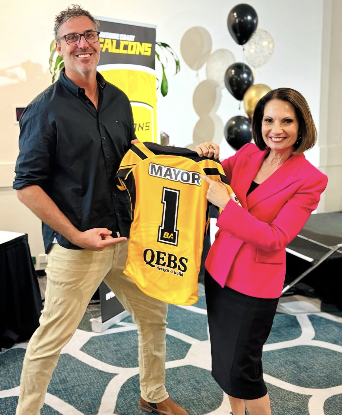 A man and a woman smiling and holding a yellow sports jersey with the number 1 and the name MAYOR, surrounded by black, white, and gold balloons, at a celebratory event.