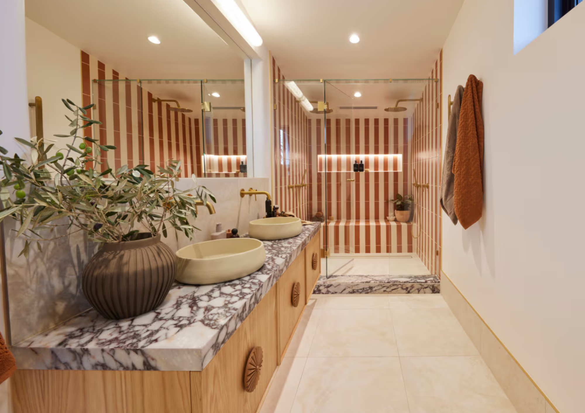 Modern bathroom with a marble countertop, two beige vessel sinks, a large mirror, and a plant in a striped ceramic pot. In the background, there is a walk-in shower with striped tile walls, glass doors, and a built-in niche with toiletries. Towel hoo