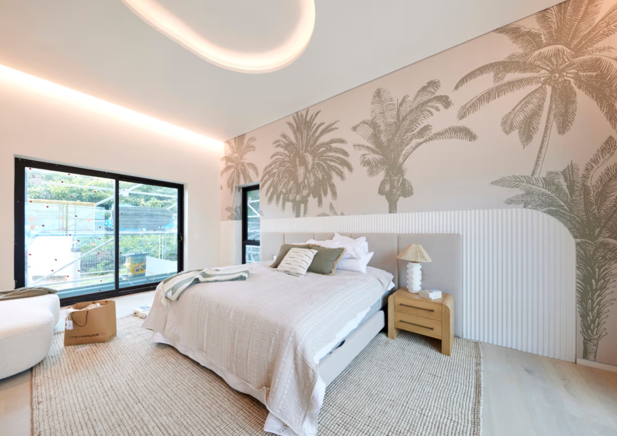 Modern bedroom with a bed, throw pillows, a wooden nightstand with a lamp, a beige rug, and large windows showing greenery outside. The wall behind the bed has a tropical palm tree mural.