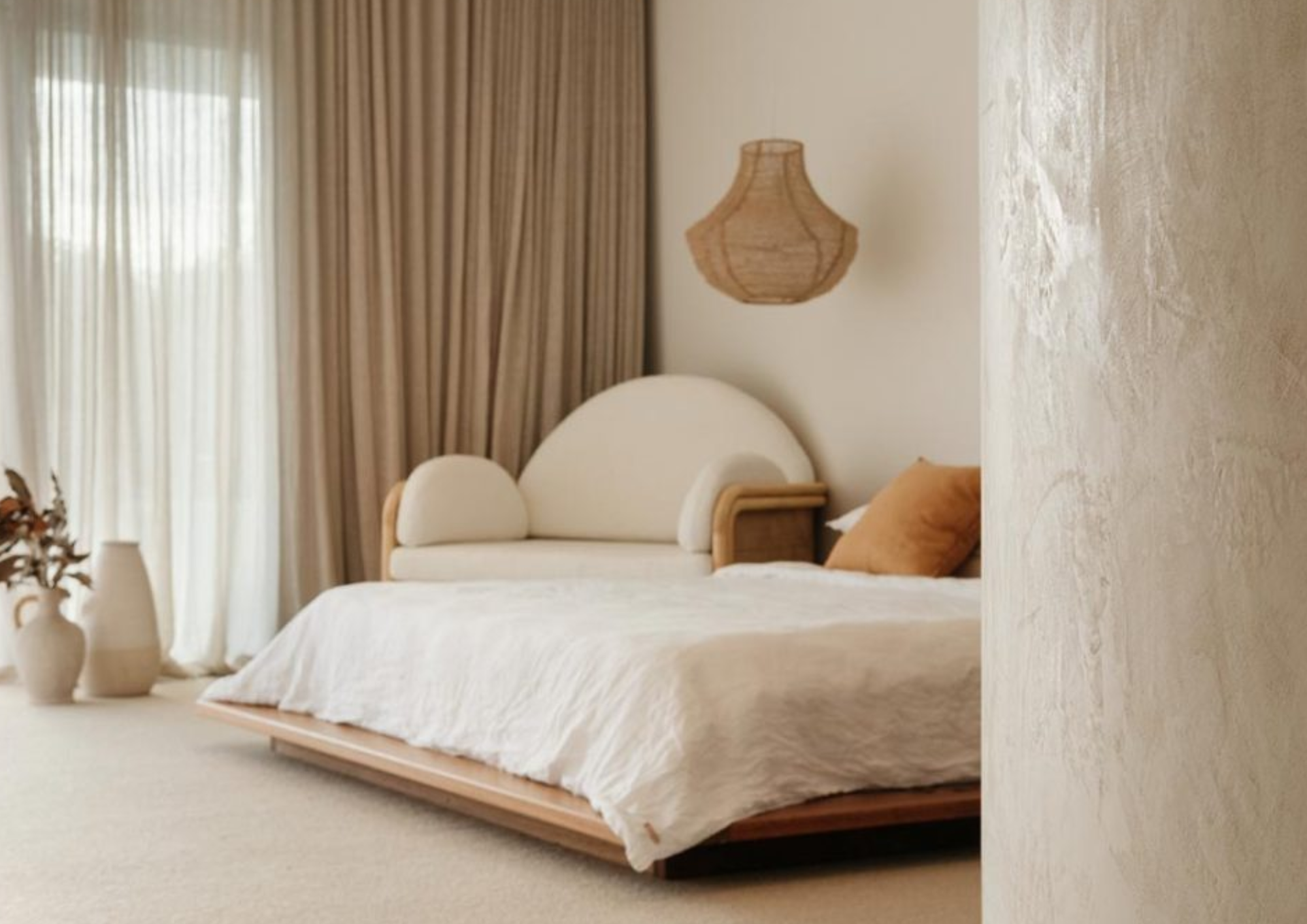 A cozy bedroom with a minimalist design featuring a bed with a curved headboard, beige bedding, and mustard-colored pillows, positioned next to a wall with a wicker wall lamp and beige curtains.