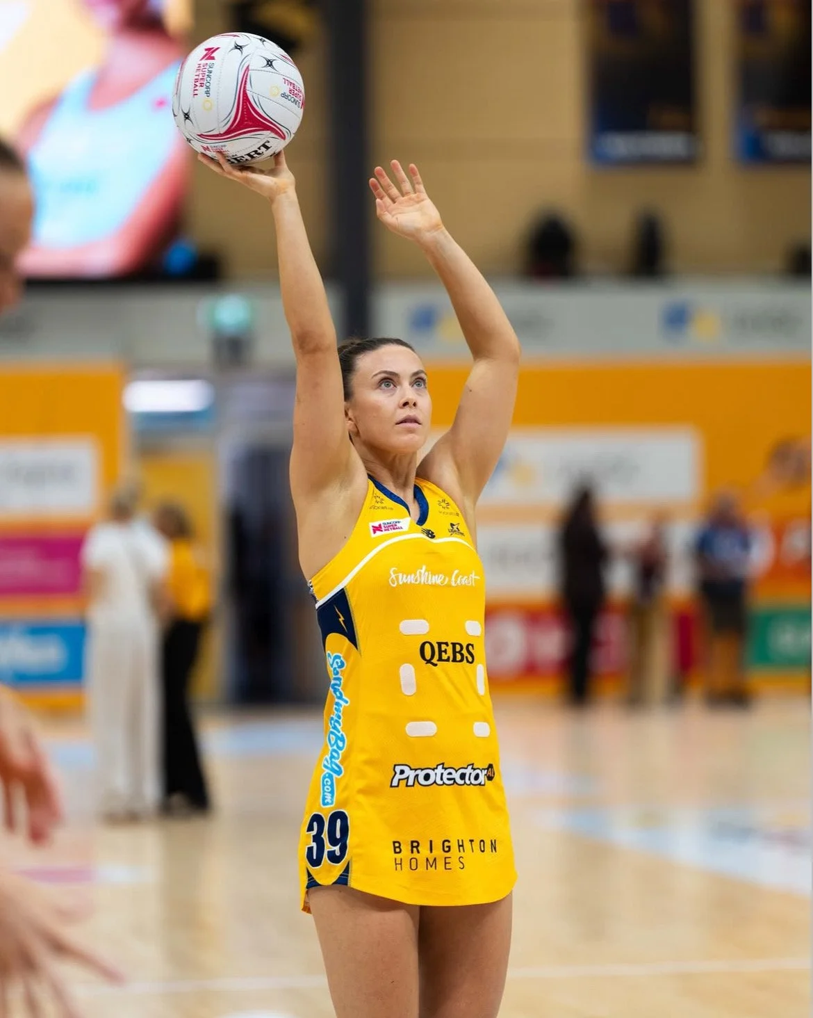 A female netball player in a yellow uniform preparing to shoot while holding a netball above her head in an indoor sports arena.