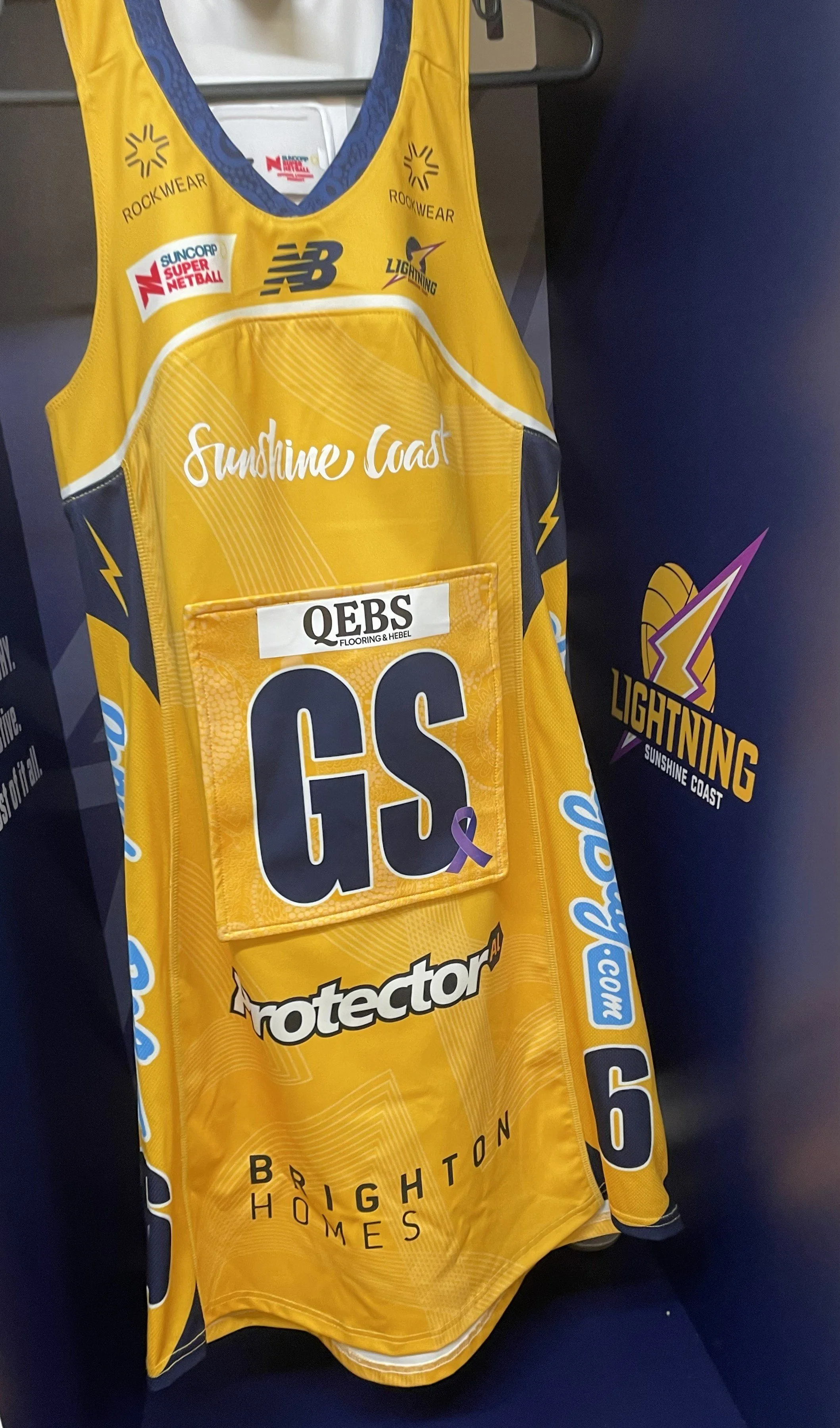 Yellow Sunshine Coast Lightning Australian Rules football jersey with team logos, sponsor patches, and a team name on the front, hanging on a hanger with a black and purple Lightning logo background.
