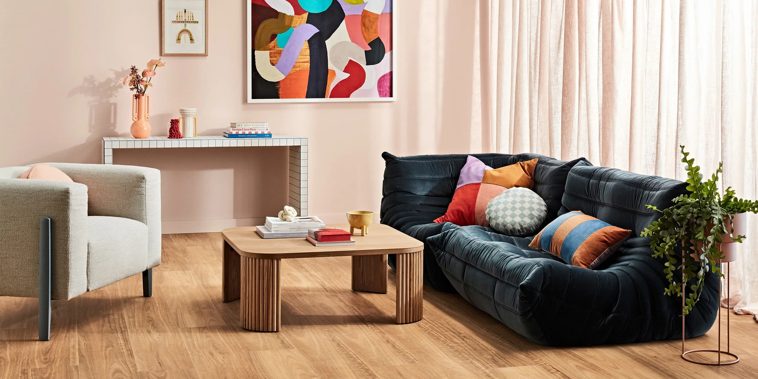 Modern living room with a black velvet sofa, colorful throw pillows, a light wooden coffee table with books and a gold bowl, a beige armchair, pink curtains, a white console table with decorative items, a colorful abstract painting, and a green plant