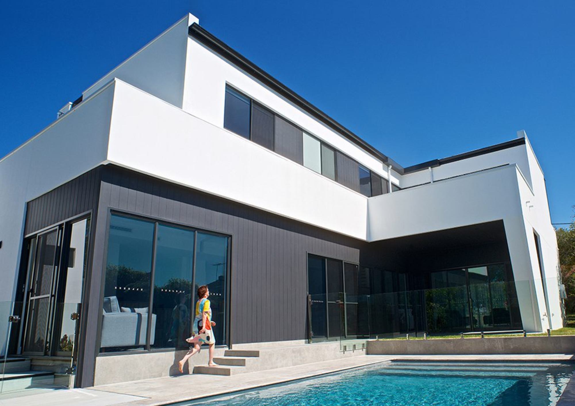 Modern multi-story house with large glass windows and a swimming pool in the foreground, under a clear blue sky.