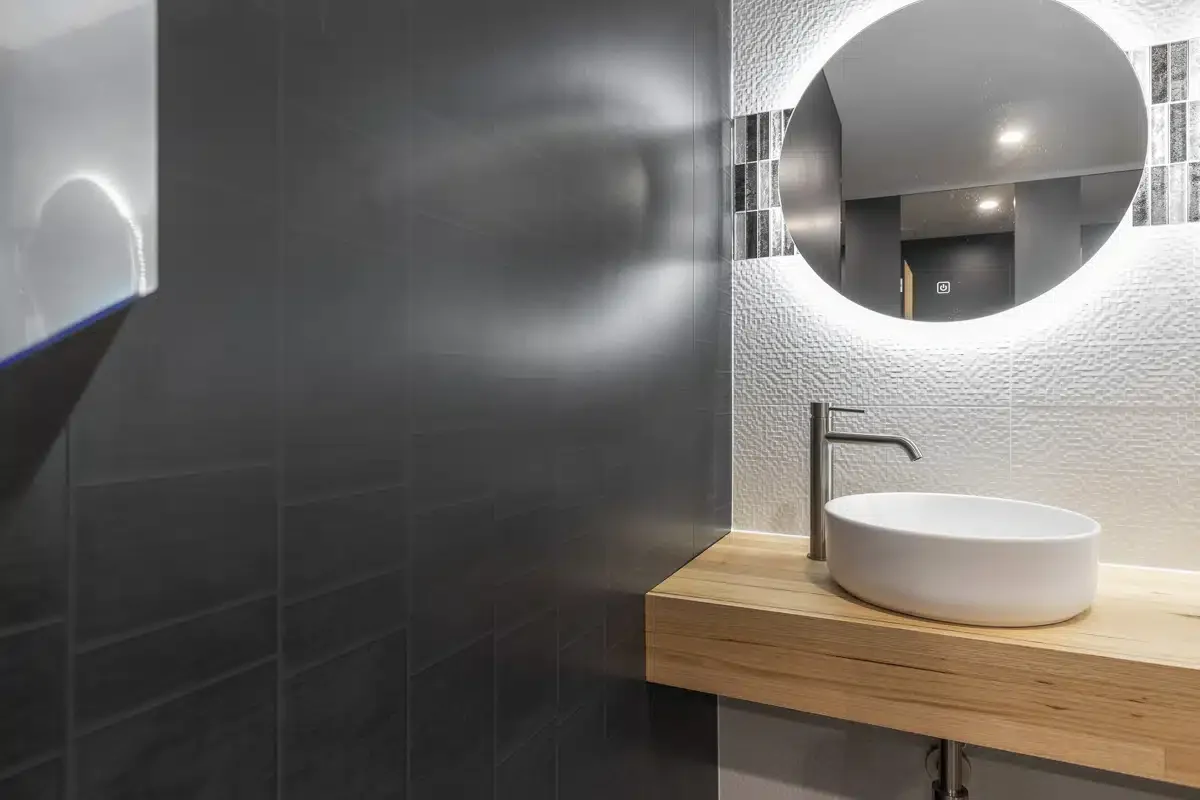 Modern bathroom with a round mirror, white textured wall tiles, a wooden countertop, and a white oval sink with a tall faucet.