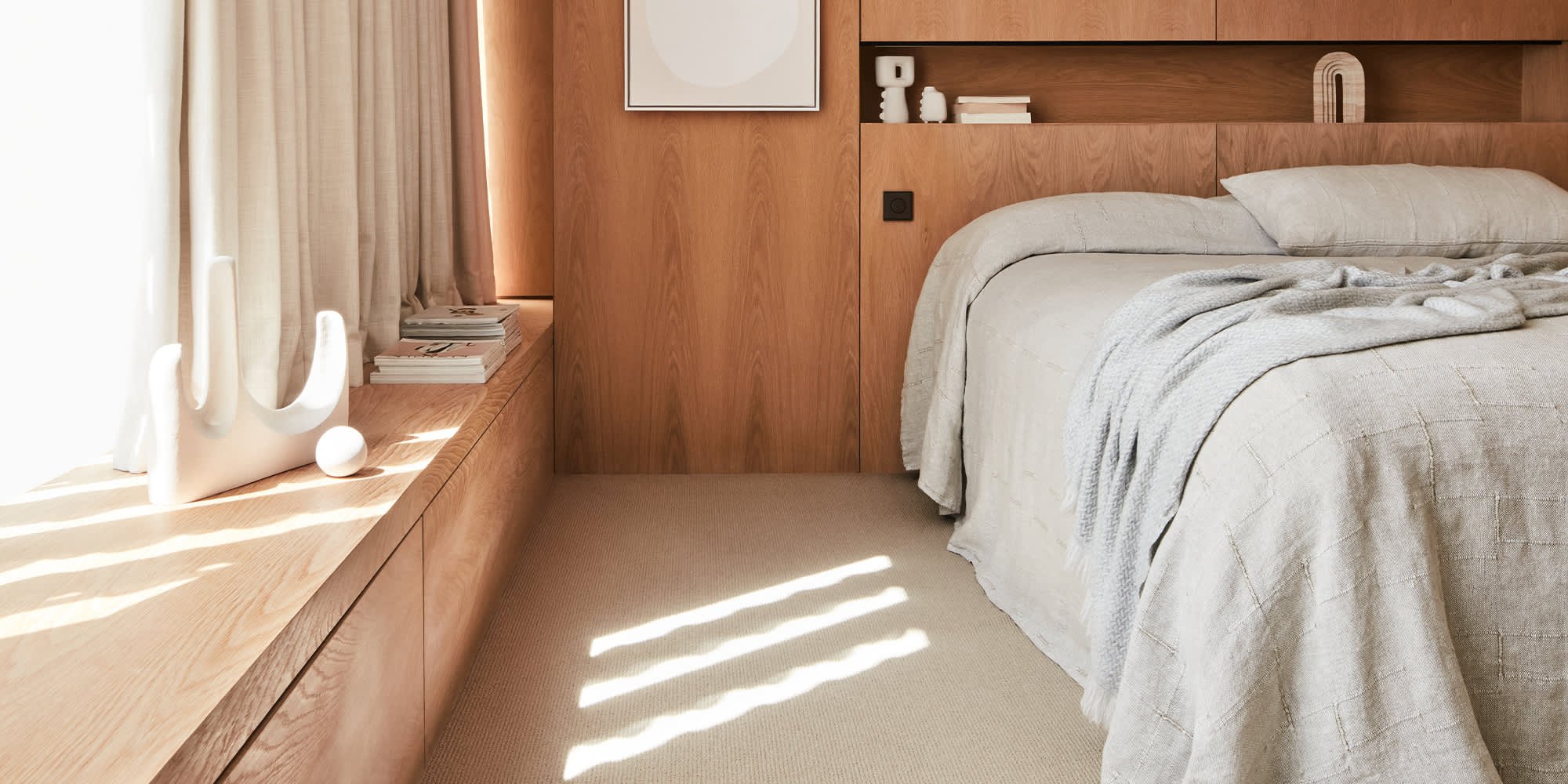 A cozy bedroom with a beige bed, a wooden headboard, and a beige curtain. Sunlight creates striped patterns on the carpeted floor, and decorative items are arranged on a wooden ledge.
