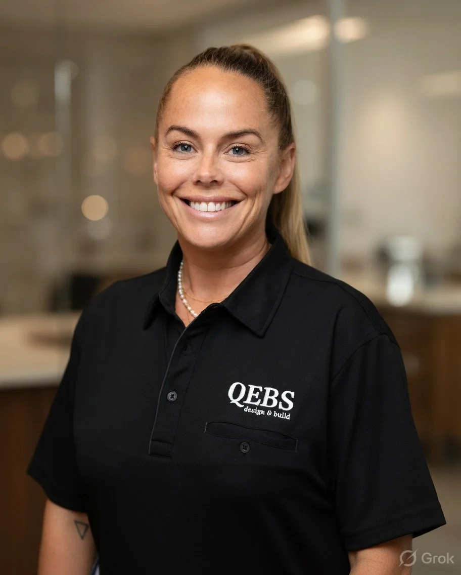 A woman with blonde hair in a ponytail, wearing a black polo shirt with 'QBS design & build' embroidered on it, smiling in an indoor setting.