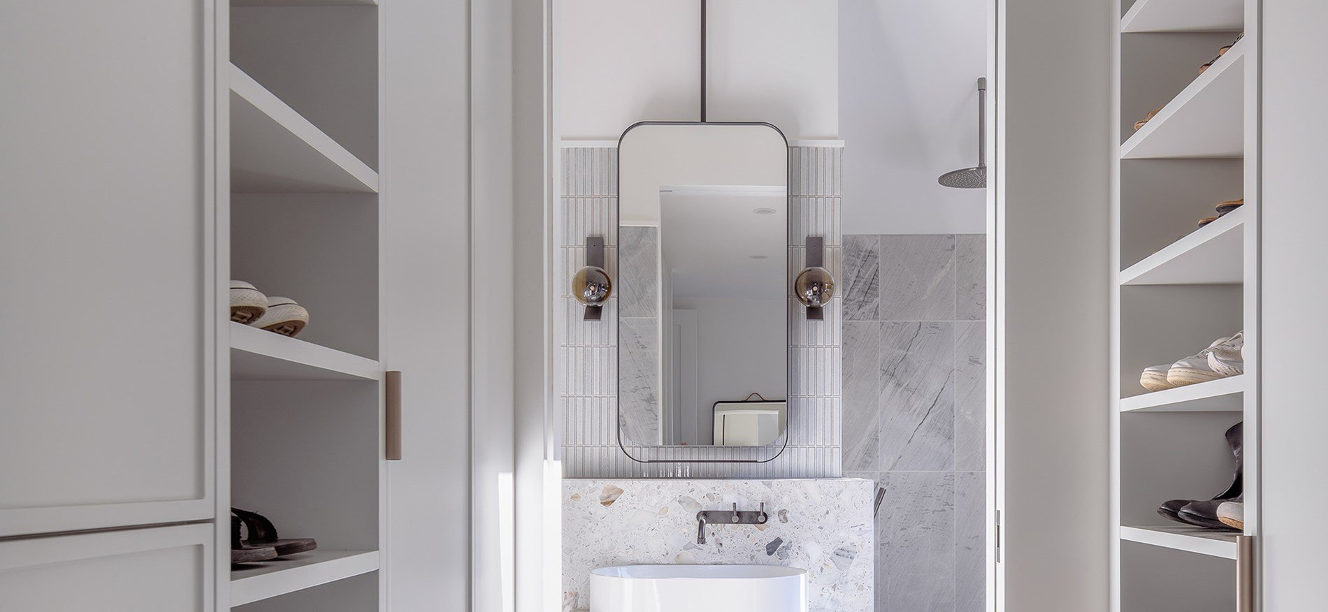 Minimalist bathroom with built-in shelves on each side, floor-to-ceiling, holding neatly arranged shoes. A tall, vertical mirror is mounted on the wall, with modern wall sconces flanking it. A rounded white bathtub is positioned below the mirror, wit