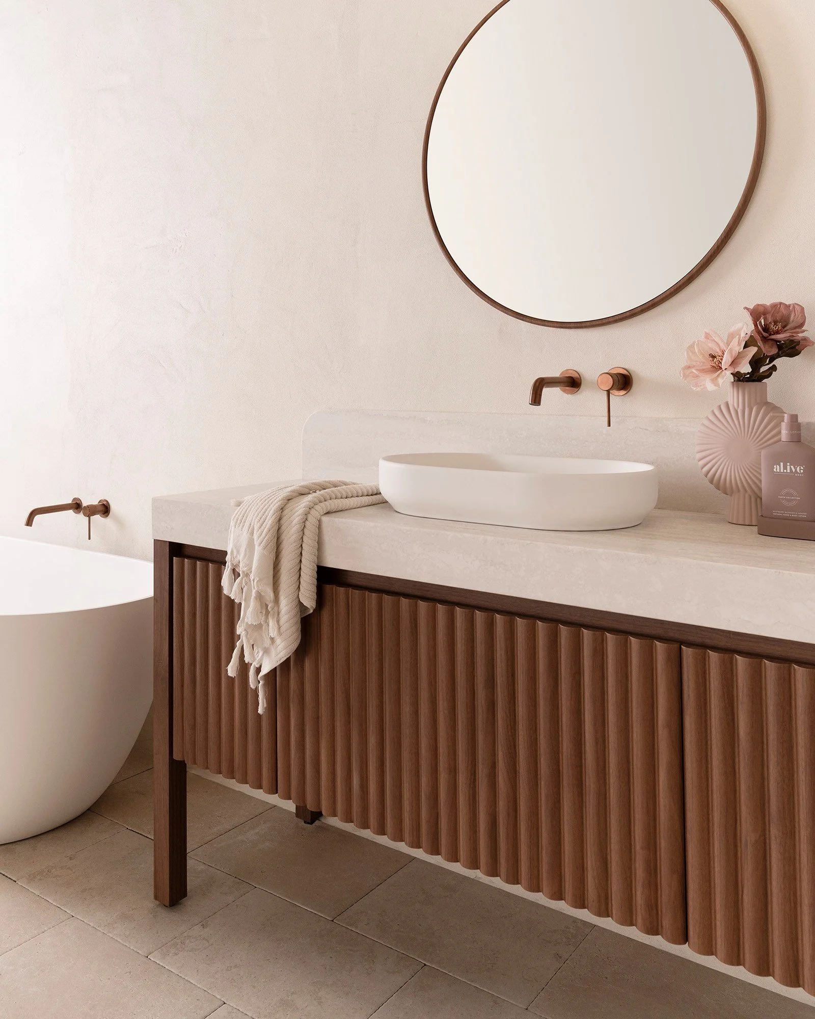 Modern bathroom with a beige countertop, white vessel sink, rose gold faucet, round mirror, pink vase with flowers, pink soap dispenser, and beige tiled floor.