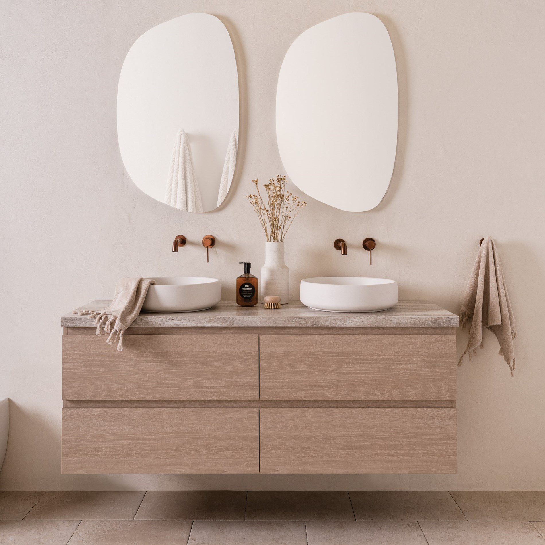 Modern bathroom vanity with two round vessel sinks, wall-mounted copper faucets, two large oval mirrors, a beige towel on the left, a towel on the right, a brown soap dispenser, a small brush, and a white vase with dried flowers.