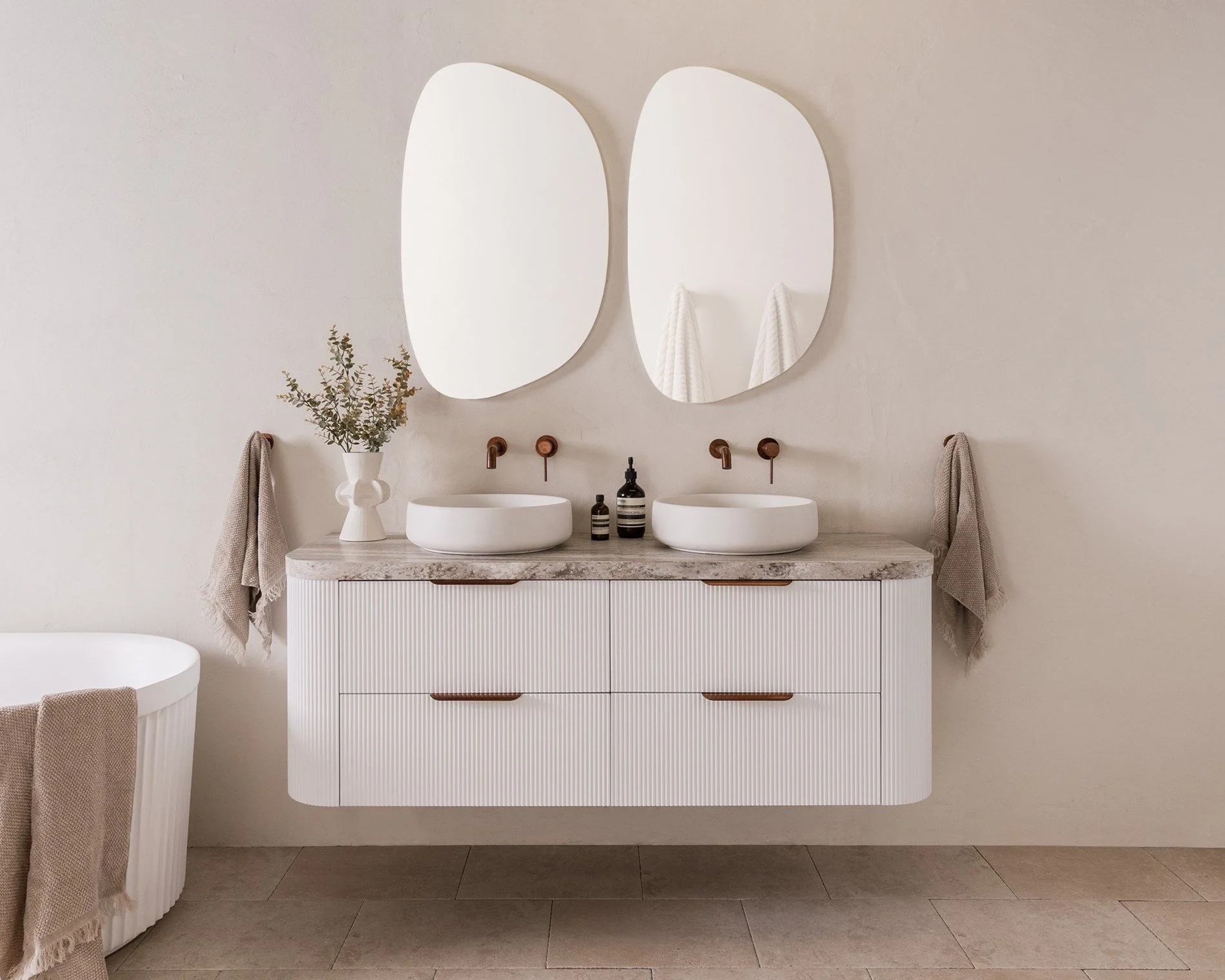 Minimalist bathroom with dual white vessel sinks, two oval mirrors, a floating vanity with natural wood accents, and beige towels.