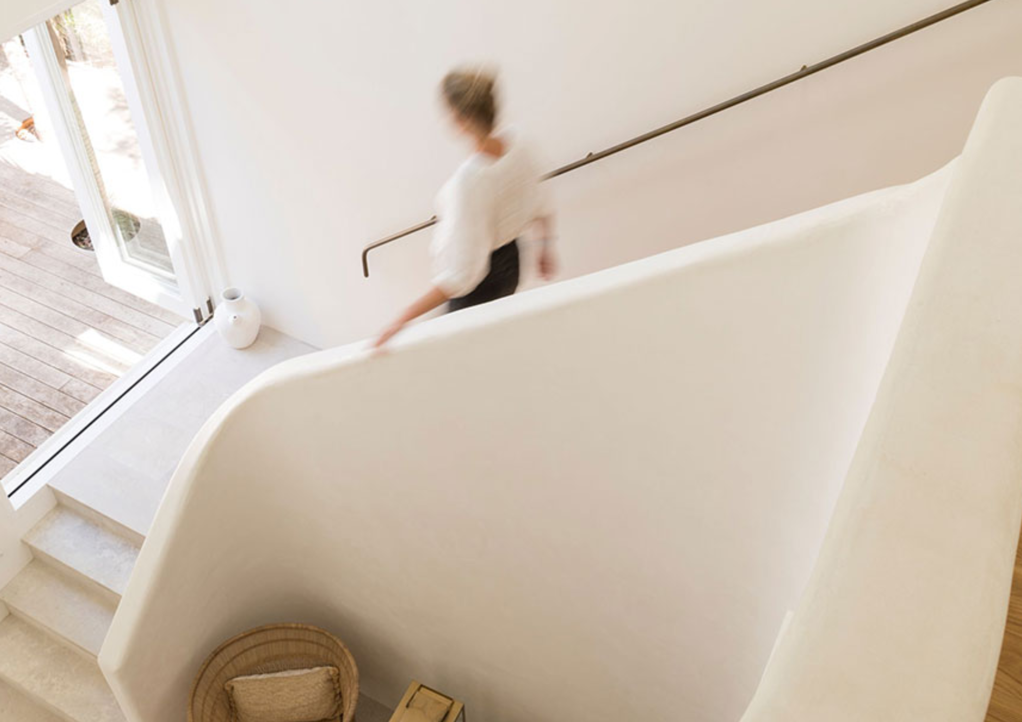 Overhead view of a woman walking up stairs near a glass door with wooden deck outside, a white vase on the floor, and a wicker basket with a blanket inside.