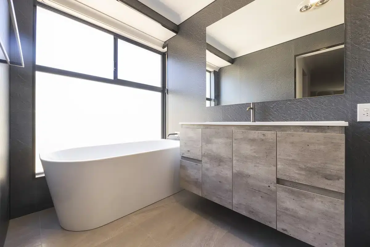 Modern bathroom featuring a freestanding bathtub near large windows, a gray textured wall, a vanity with wooden cabinets, a white countertop, a mirror, and a ceiling with built-in lighting.