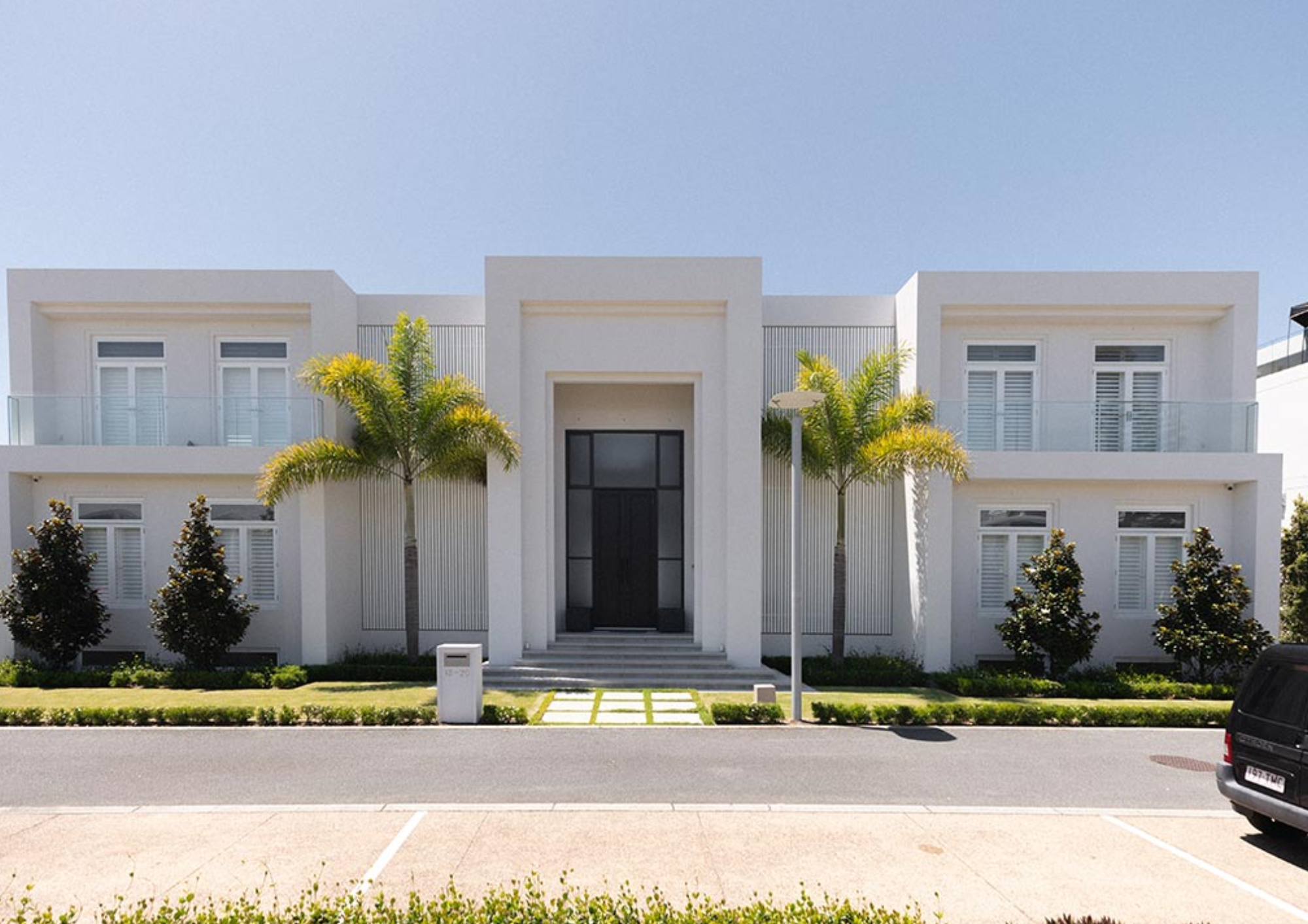 Modern white two-story residential building with glass balconies and palm trees in front.