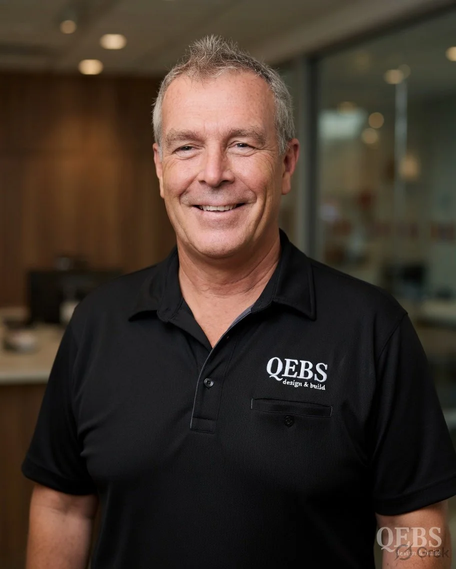 Smiling man wearing a black polo shirt with QBS logo, standing indoors with blurred background.