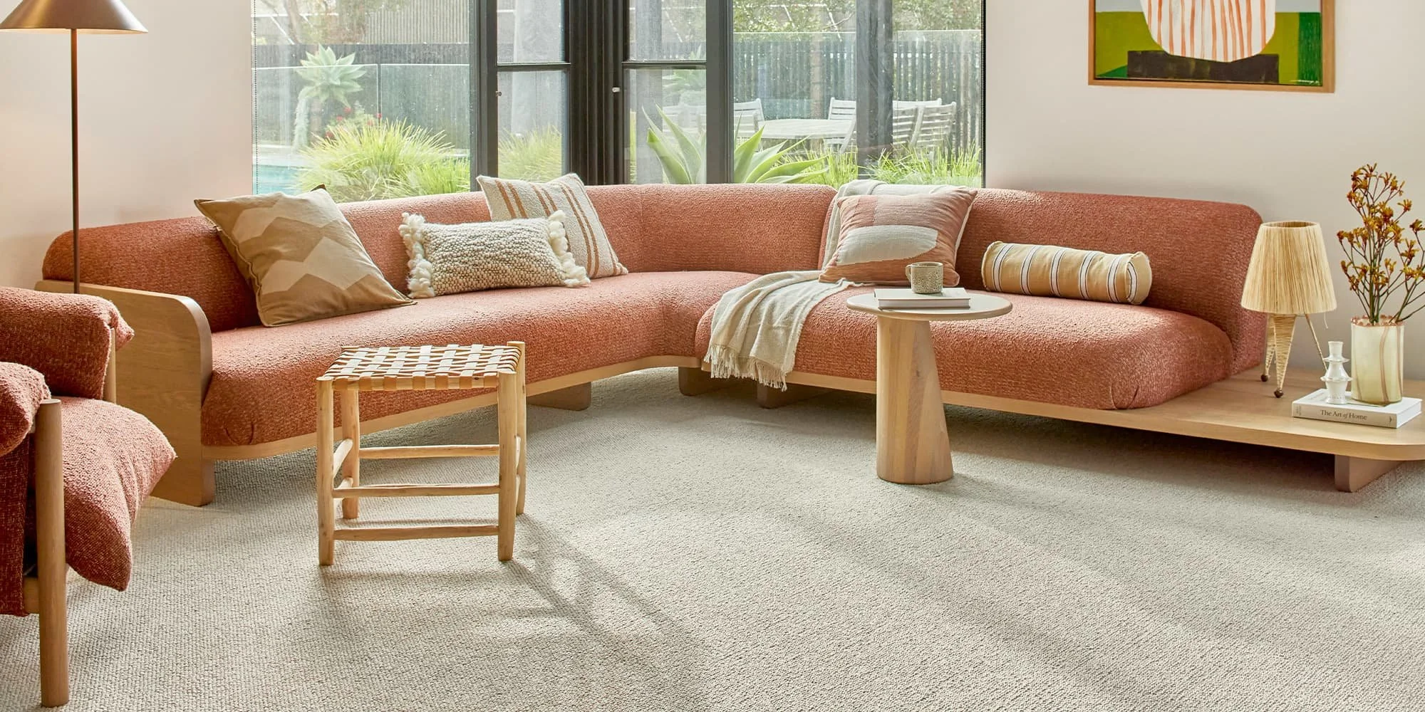 Cozy living room with a large pink sectional sofa, decorative pillows, a small wooden side table, and a floor lamp. Large windows with a view of an outdoor patio and garden.