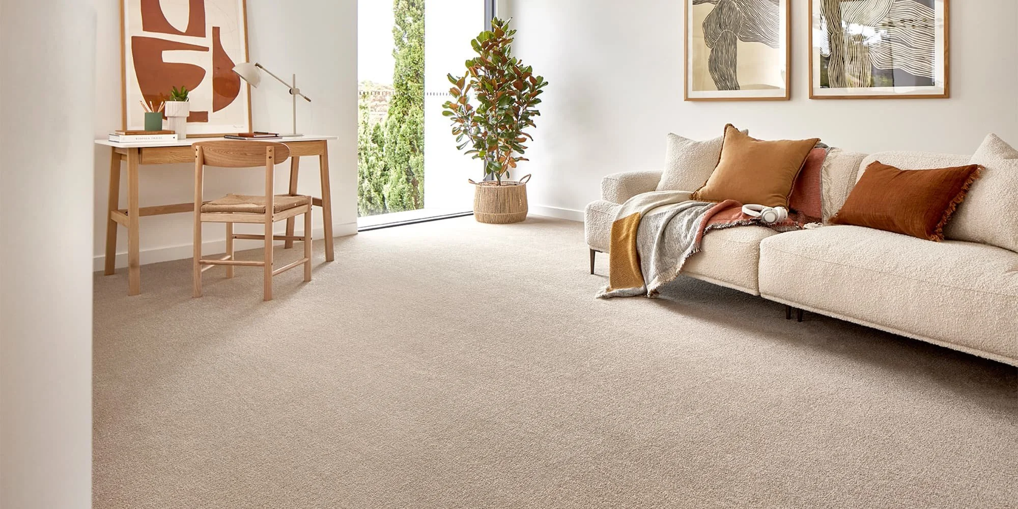 Living room with beige carpet, cream sofa with brown and cream cushions, a throw blanket. A light wood desk with a chair, a white table lamp, books, and a small plant. Large window with greenery outside, a tall houseplant in a woven basket, and three