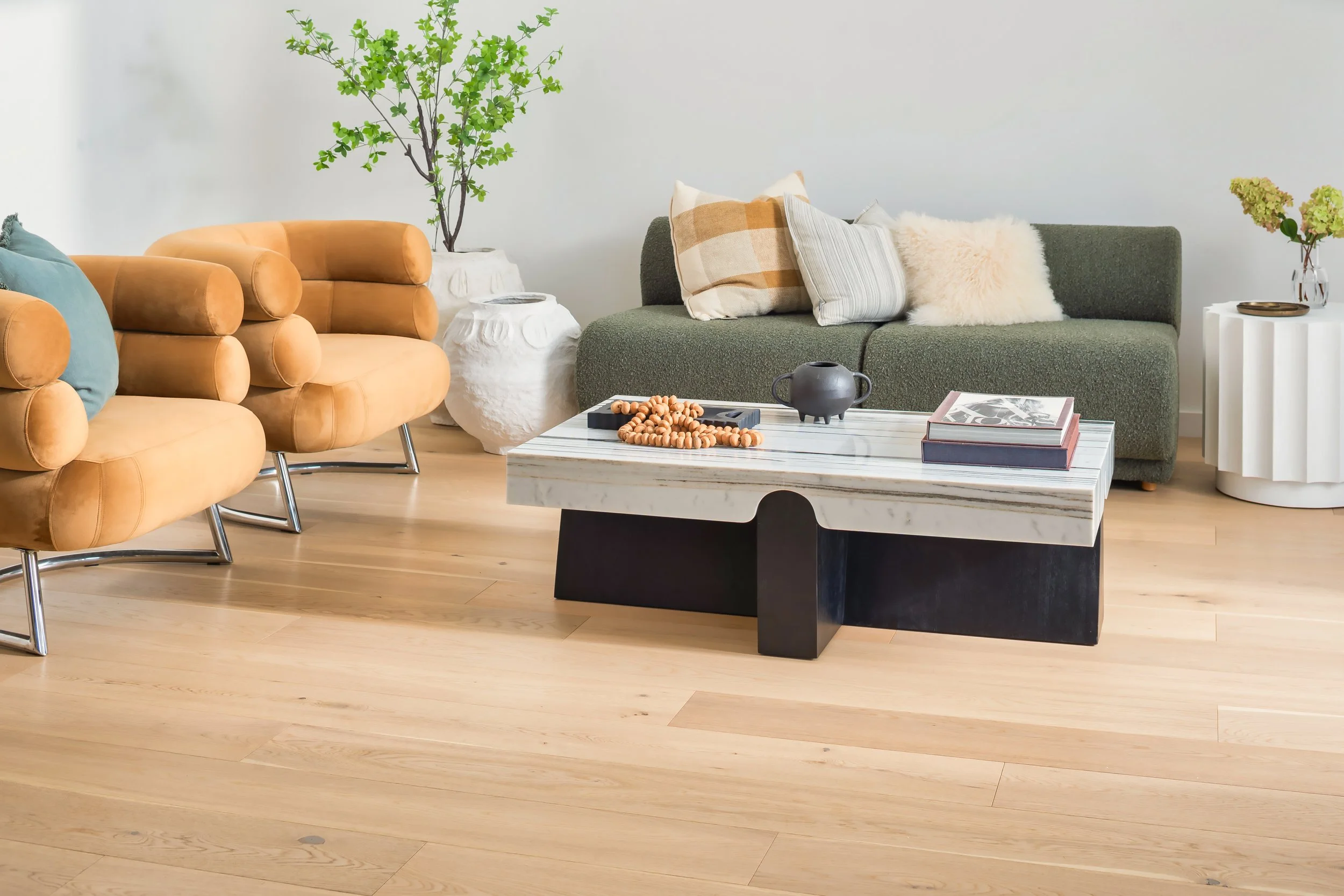 Living room with two orange velvet armchairs, a green fabric sofa with multiple pillows, a white and black marble coffee table with books, a black teapot, and a wooden beaded necklace. Decor includes a large green plant, white textured side tables wi