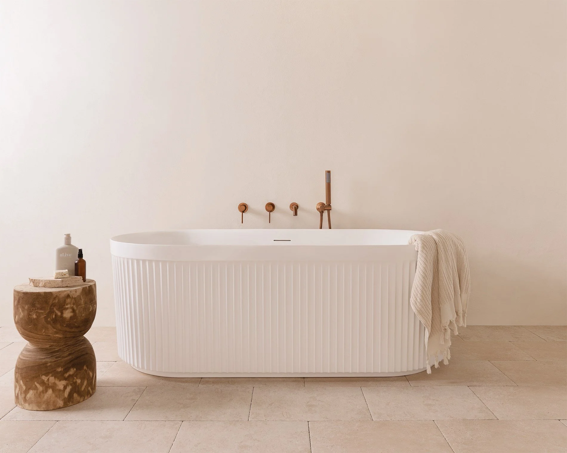 A modern white bathtub with vertical ribbed design, a beige towel draped on the side, and a small, rustic wooden side table with bottles and soap on it. The bathroom features beige tile flooring and minimalistic fixtures.