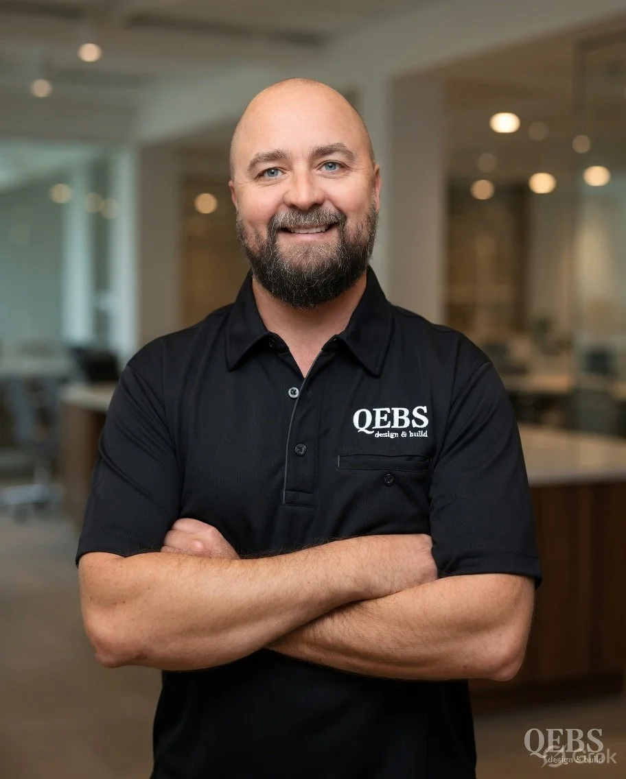 Older man with a beard and bald head wearing a black polo shirt with 'QEBS design & build' logo, standing indoors with arms crossed, smiling.