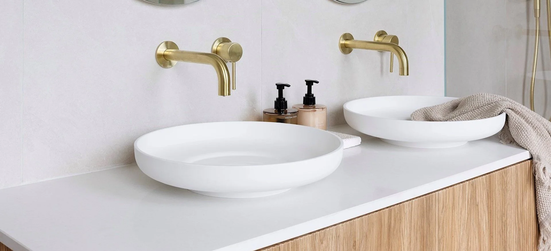 Bathroom double vanity with two white oval vessel sinks, gold wall-mounted faucets, soap dispensers, a folded towel, and a beige textured cloth, with a light wood cabinet and a neutral-colored tiled wall.
