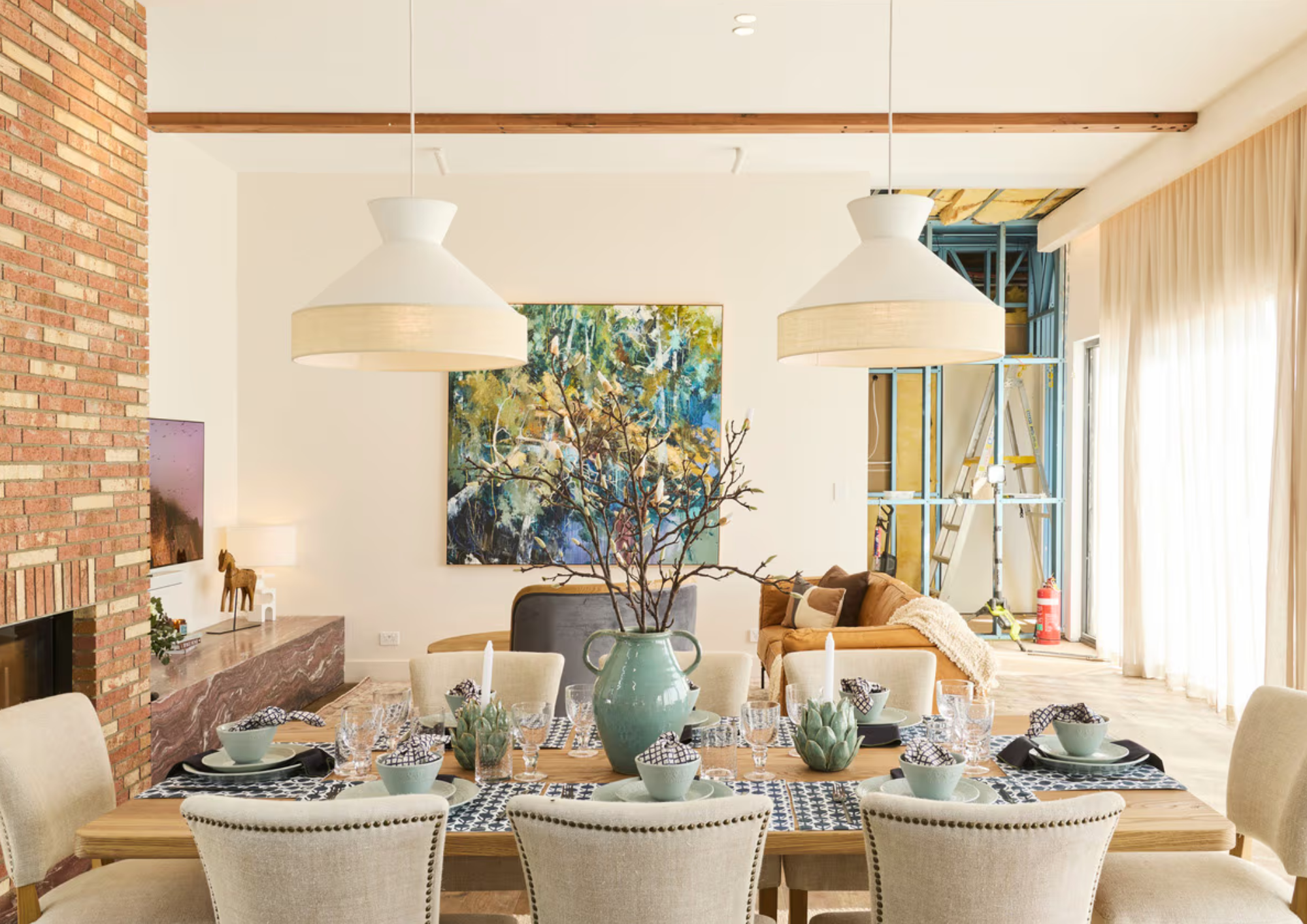A dining room with a wooden table set with bowls, glasses, and utensils, surrounded by cream-colored chairs. A large green vase with leafless branches is at the center of the table. In the background, there is a living space with a brown sofa, a colo