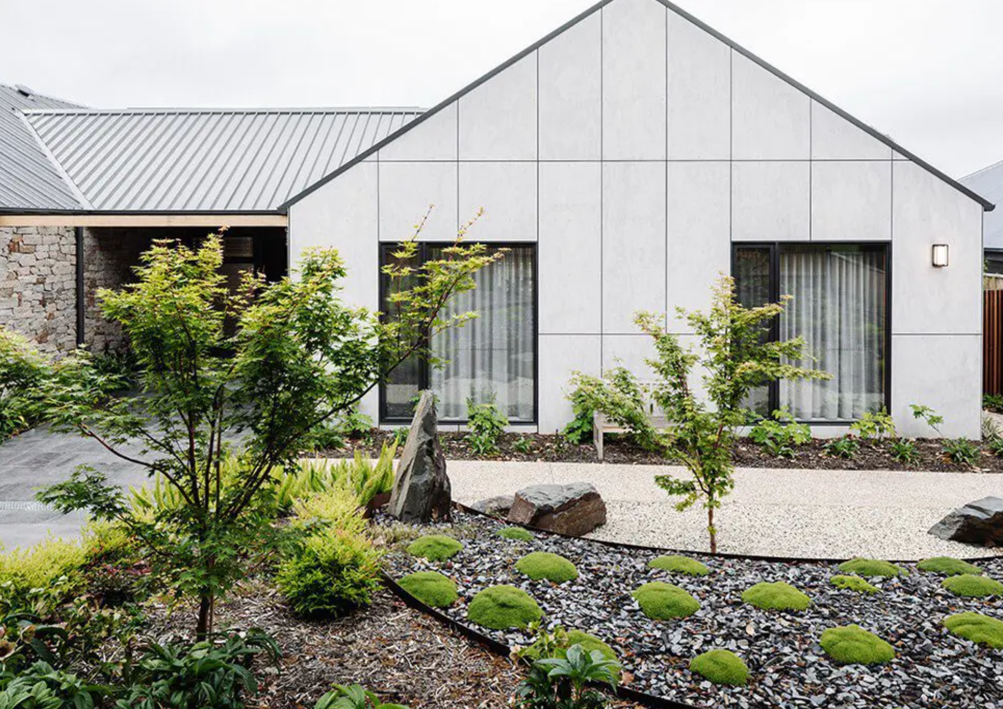 Modern house with a white exterior, large windows with curtains, gabled roof, and landscaped front yard with small trees, rocks, and gravel path.