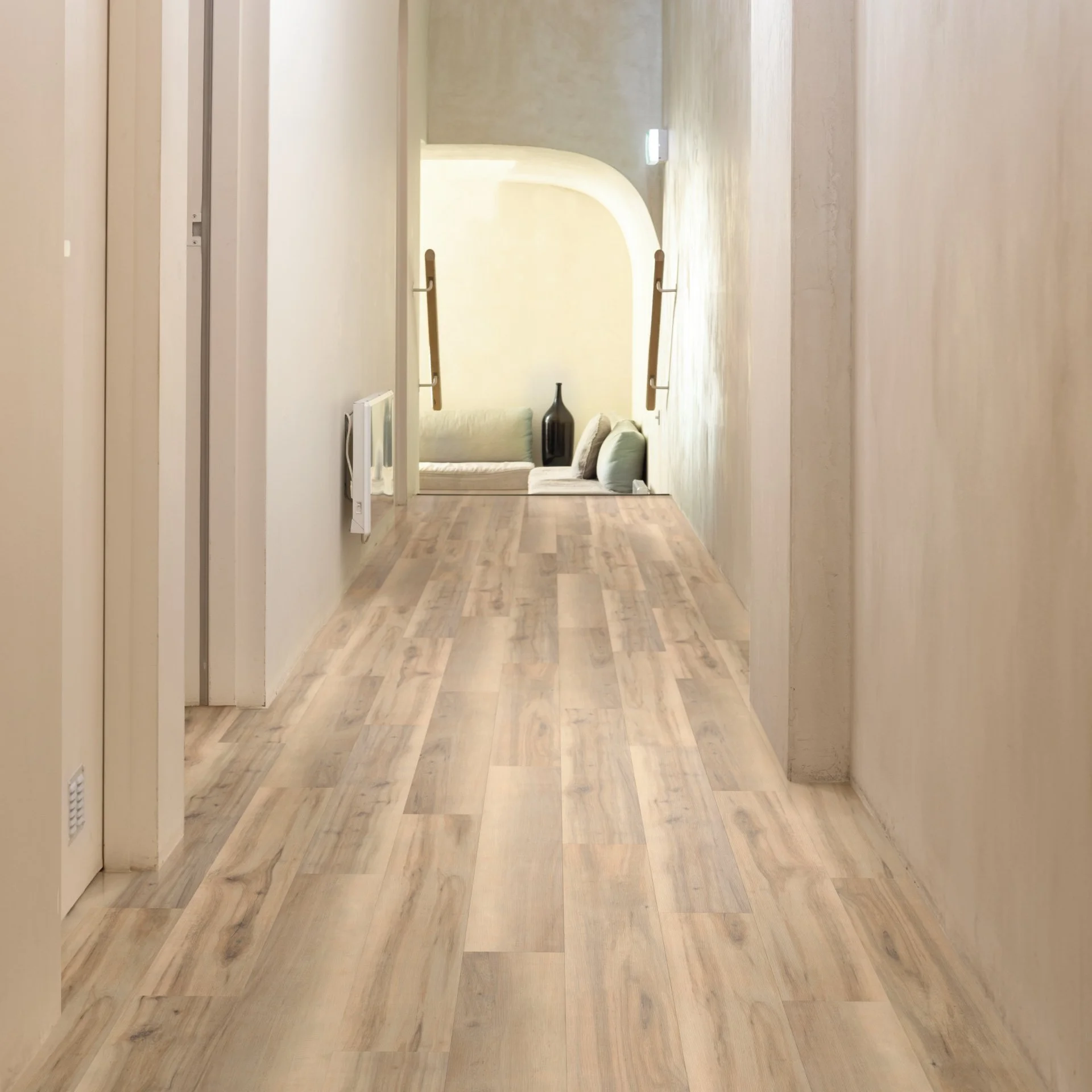 A hallway with light-colored wooden flooring leading to a small sitting area with a couch and a black vase.