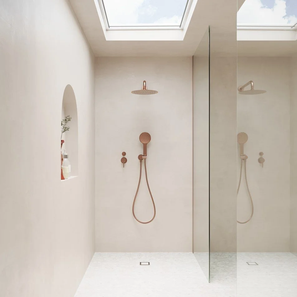A minimalist shower with beige walls, a skylight, and a large mirror reflecting a copper showerhead and handheld wand, with a built-in shelf holding toiletries.