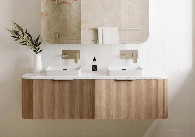 Modern bathroom with a floating wooden vanity with two white vessel sinks, a large mirror, wall-mounted faucets, a bottle of hand soap, and a vase with green leaves.