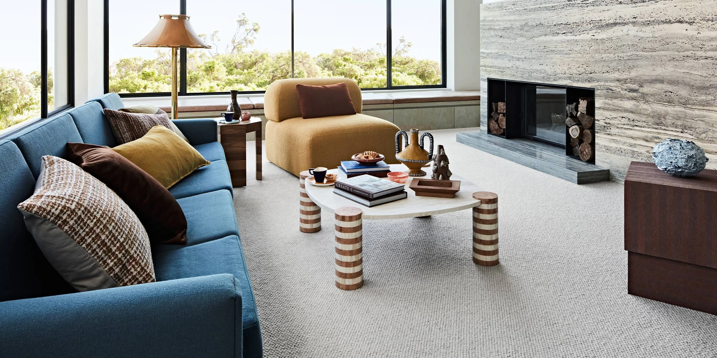 Living room with a blue sofa, yellow and brown cushions, a beige armchair, a white coffee table with books and decorative items, large windows showing greenery outside, a modern fireplace with firewood, and a stone sculpture on a wooden side table.