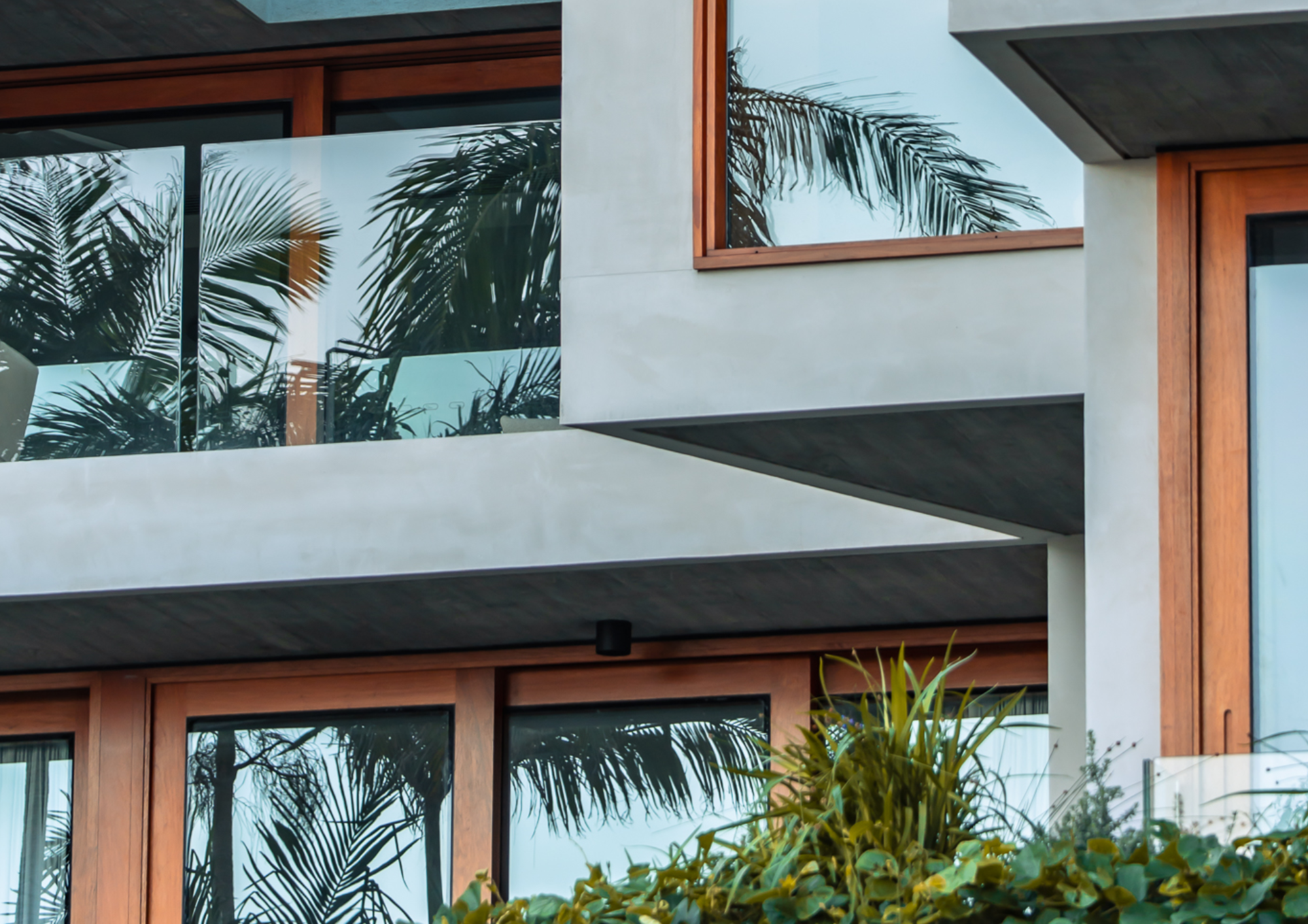 Close-up of a modern building with large glass windows, wooden frames, concrete elements, and tropical plants reflected in the glass.