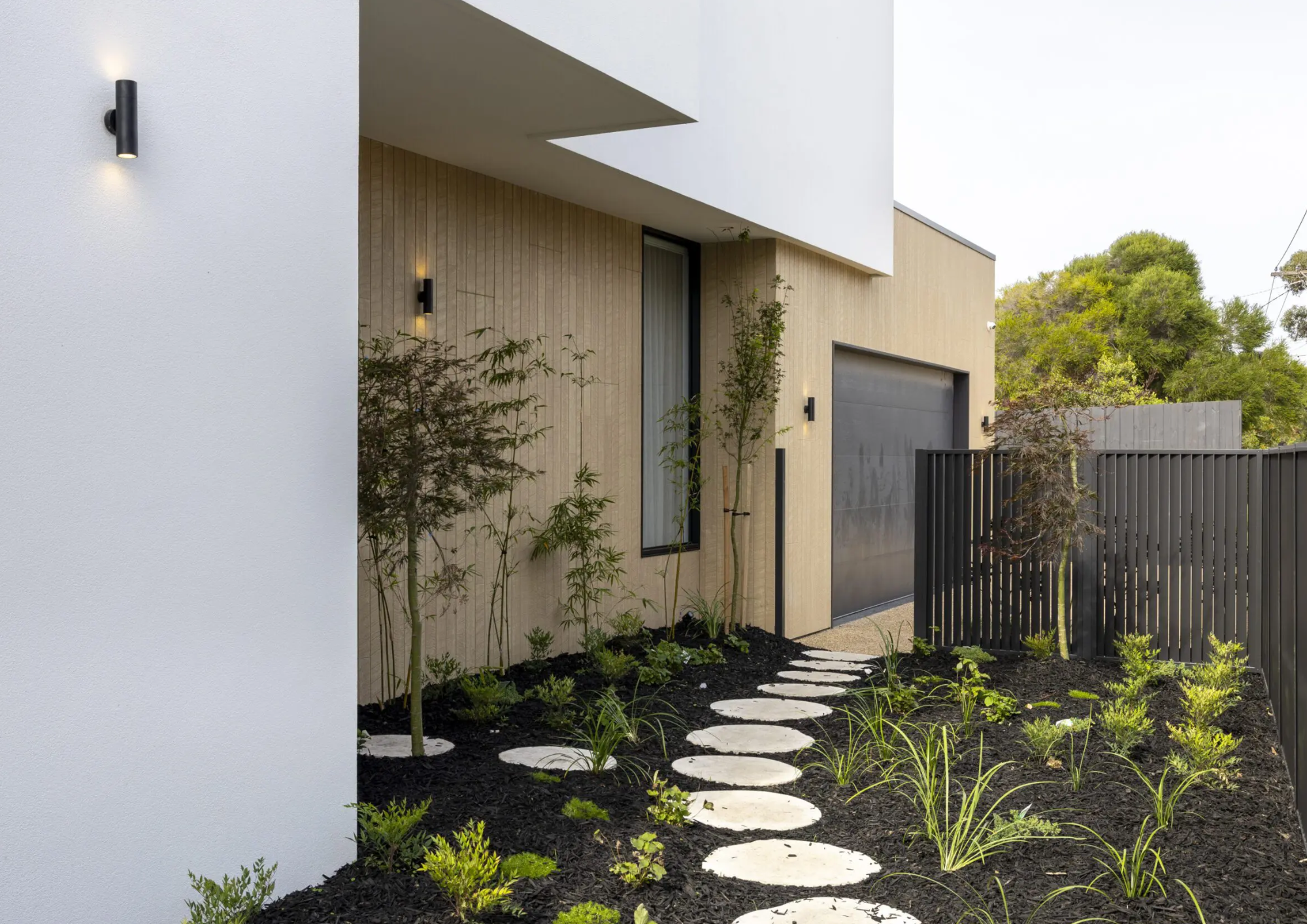 Modern house with a white and beige exterior, black garage door, and a small front garden with a pathway made of large white stepping stones, various plants, and trees, surrounded by a black fence.