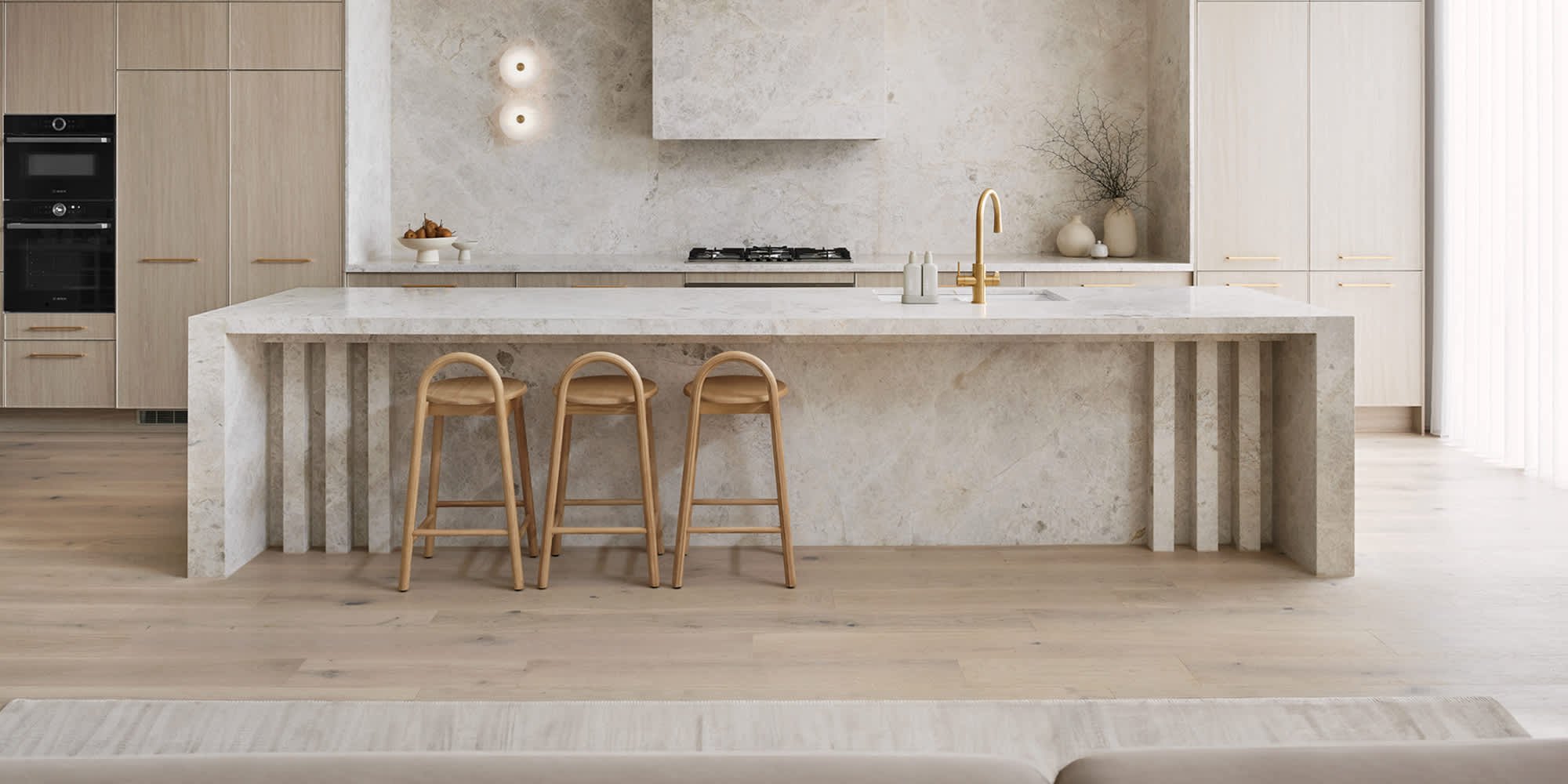 Modern kitchen with beige stone island and matching cabinets, three wooden barstools, built-in oven and microwave, gold faucet, white and beige decor, and light wooden flooring.