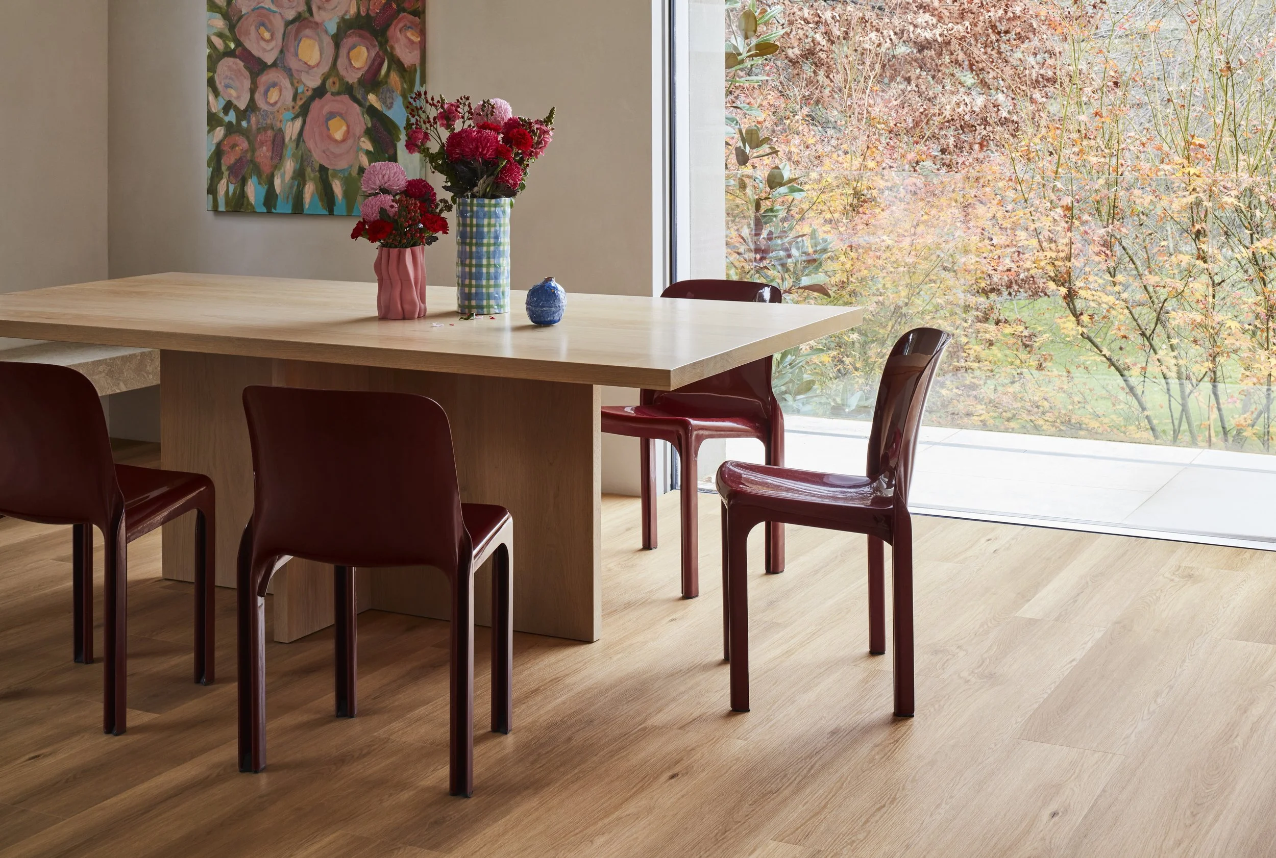 Dining room with a wooden table, red chairs, colorful flowers in vases, and a large window showing trees with fall foliage.