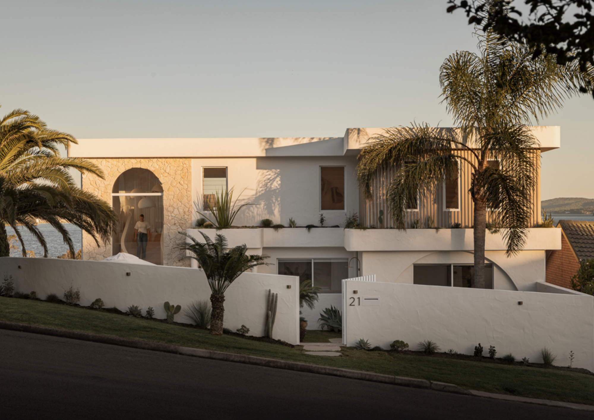 Modern white two-story house with rounded and angular design elements, surrounded by palm trees and a white wall with house number 21, views of water in the background during sunset.