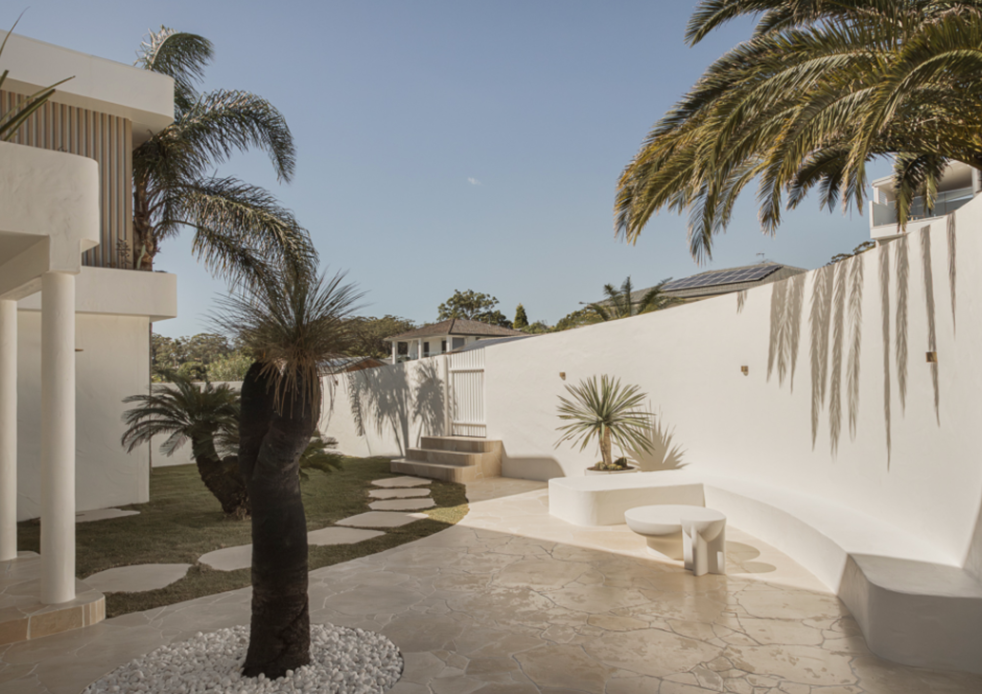 A modern outdoor patio with white curved seating, palm trees, and a pathway leading to a gate, enclosed by a white wall.
