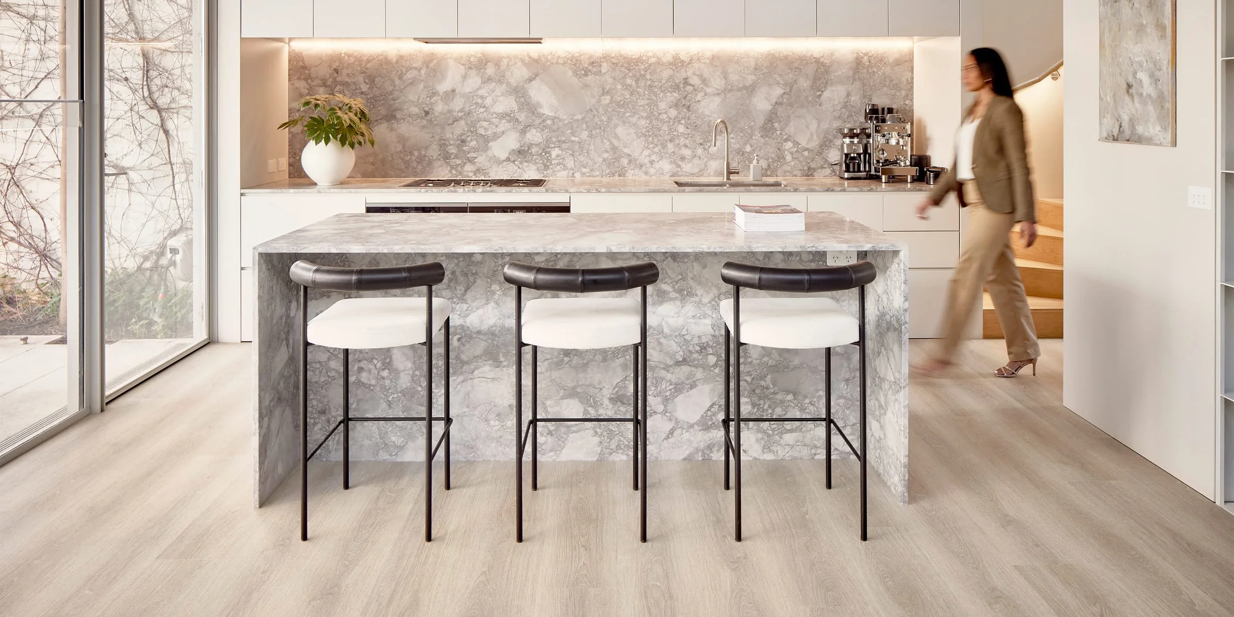Modern kitchen with a marble island, three barstools, a potted plant, and a woman walking upstairs