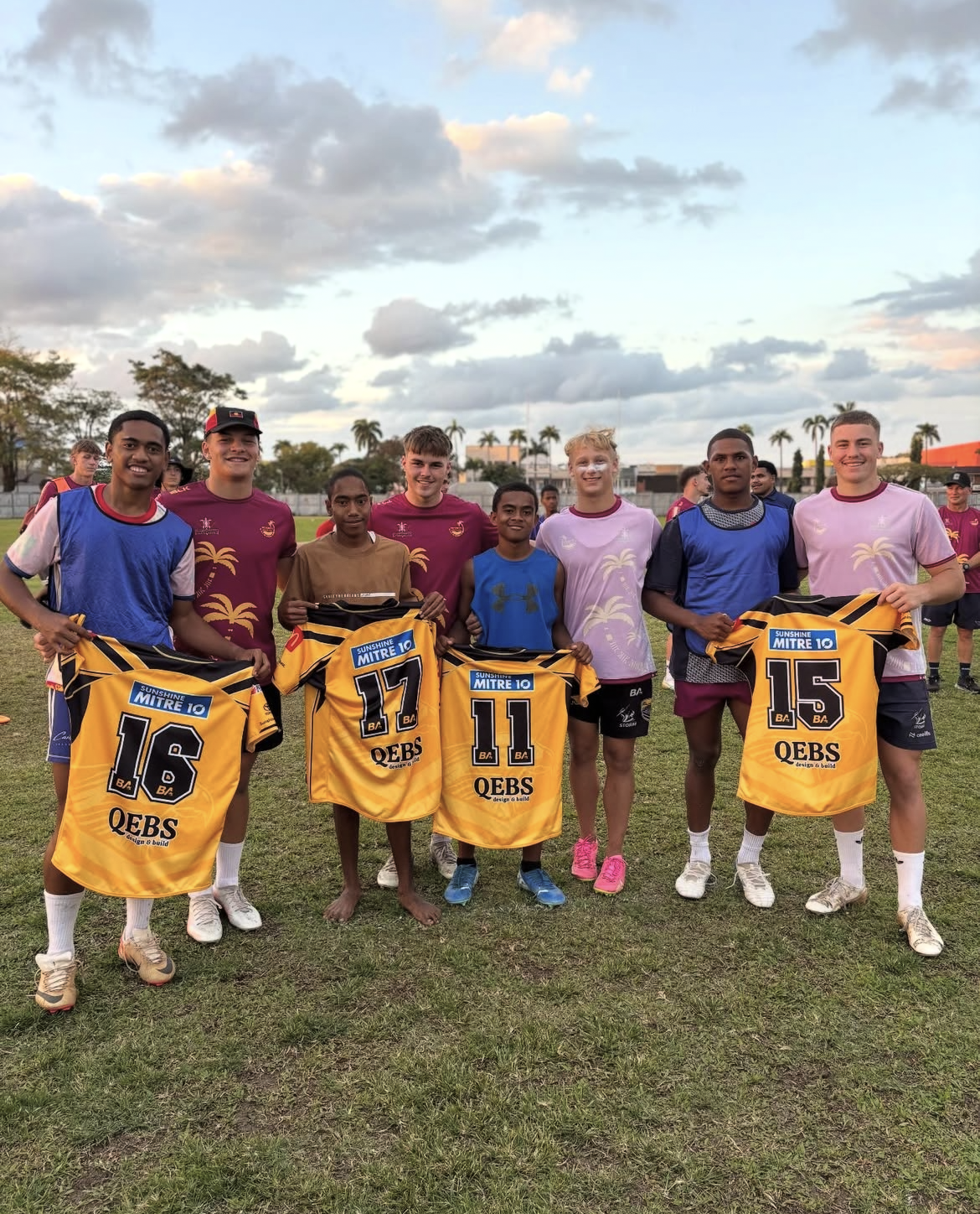 A group of young male soccer players standing on a grassy field, holding yellow jerseys with black numbers. They are smiling, with some wearing different colored jerseys, and there are palm trees and buildings in the background.