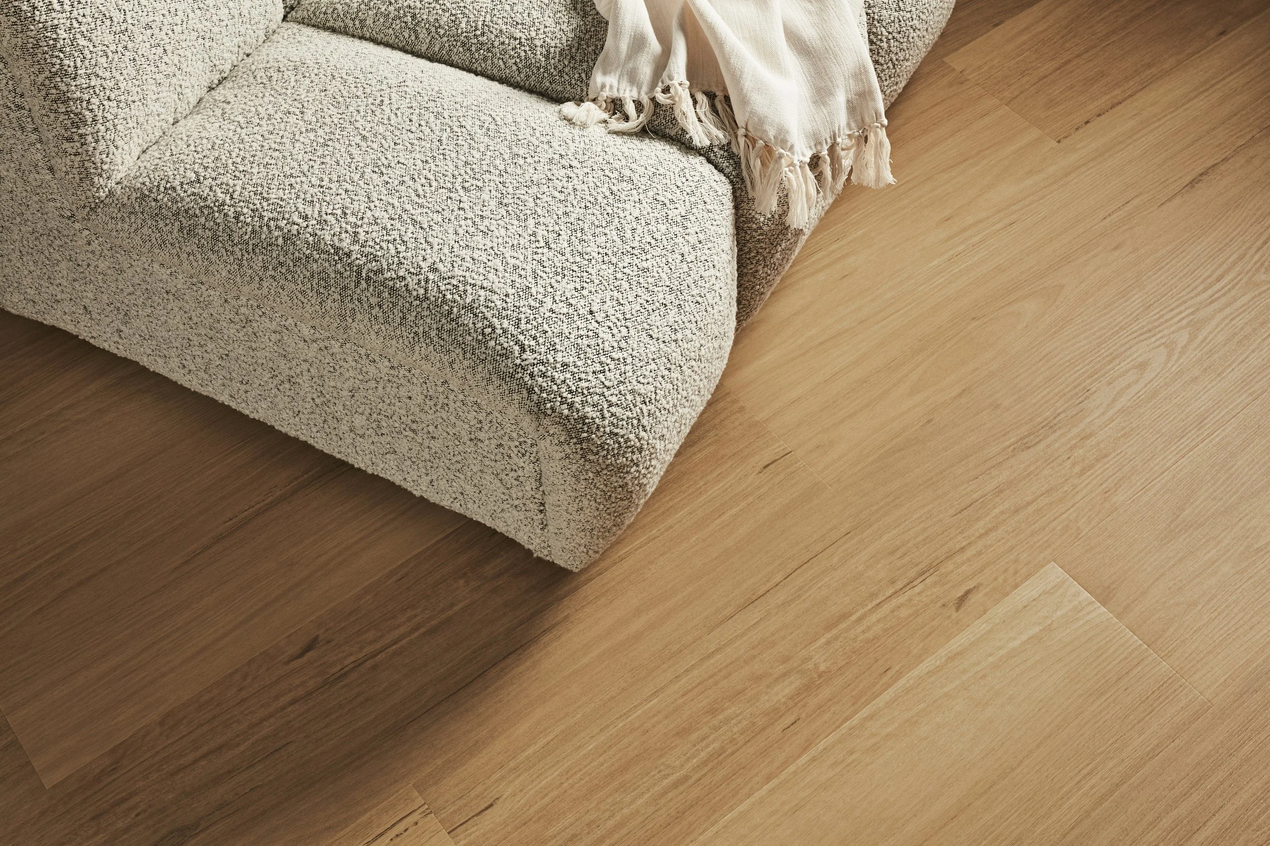 Close-up of a beige fabric sofa with a textured pattern, partially covered by a white fringed throw blanket, on a light wood floor.