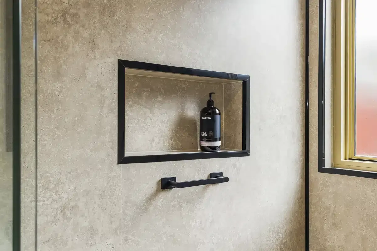 A shower with beige textured tile walls, a built-in black-framed shelf holding a black body wash bottle, and a black towel bar below the shelf. There is a window with a beige frame and frosted glass on the right side.