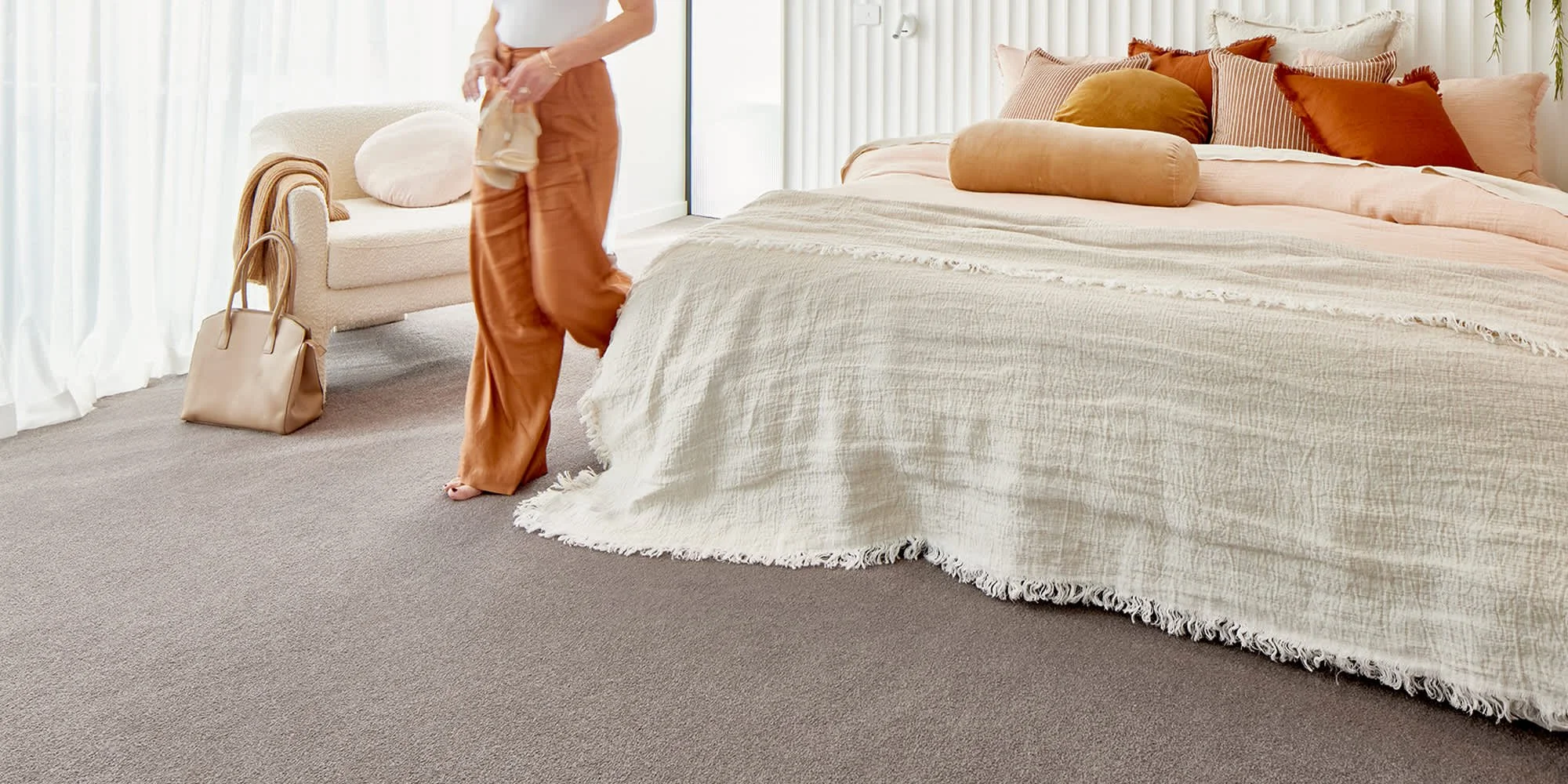 Person walking in a bedroom with a large bed, layered blankets, and multiple throw pillows, with a sitting area and a bag nearby.