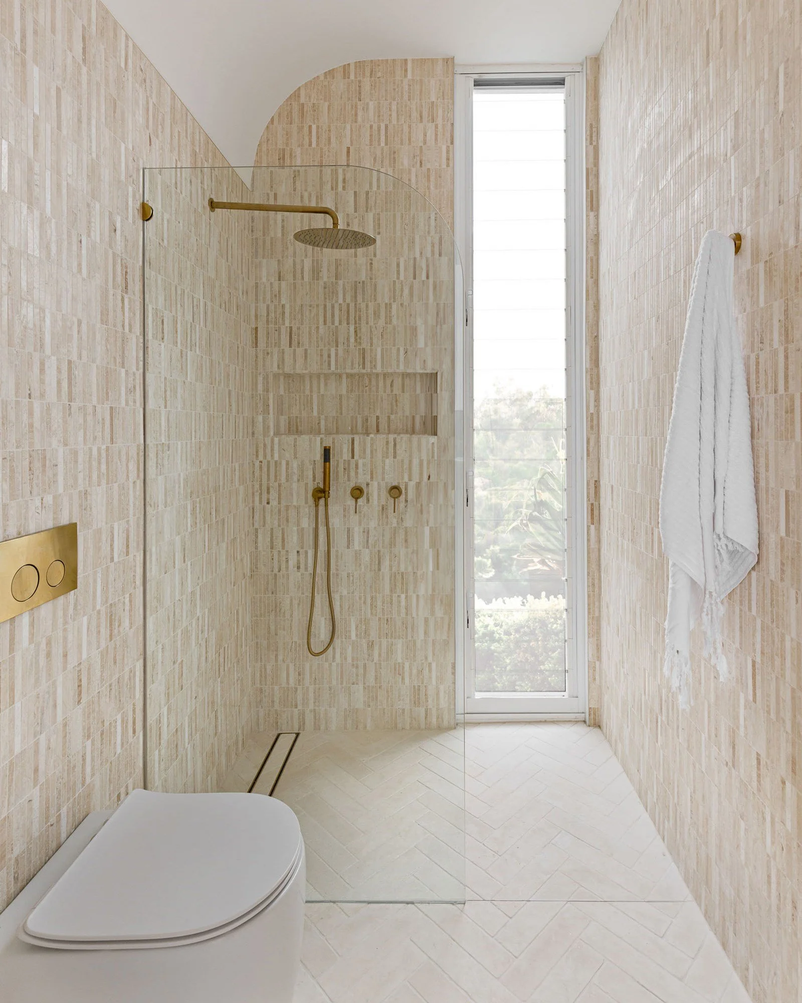 A modern bathroom with a walk-in shower, beige tile walls, a glass shower partition, a window with a view outside, a wall-mounted white toilet, and a white towel hanging on the wall.