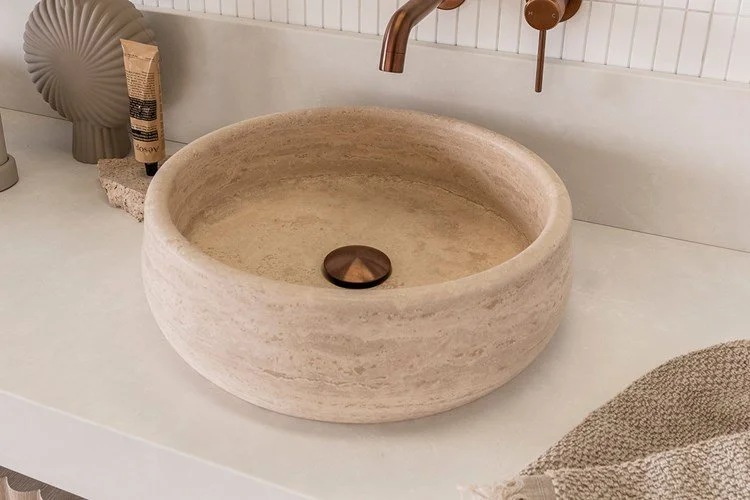 A round stone bathroom sink with a bronze drain, set on a white countertop. Nearby are decorative items including a textured ceramic vase, a tube of lotion, a stone coaster, and a woven towel.