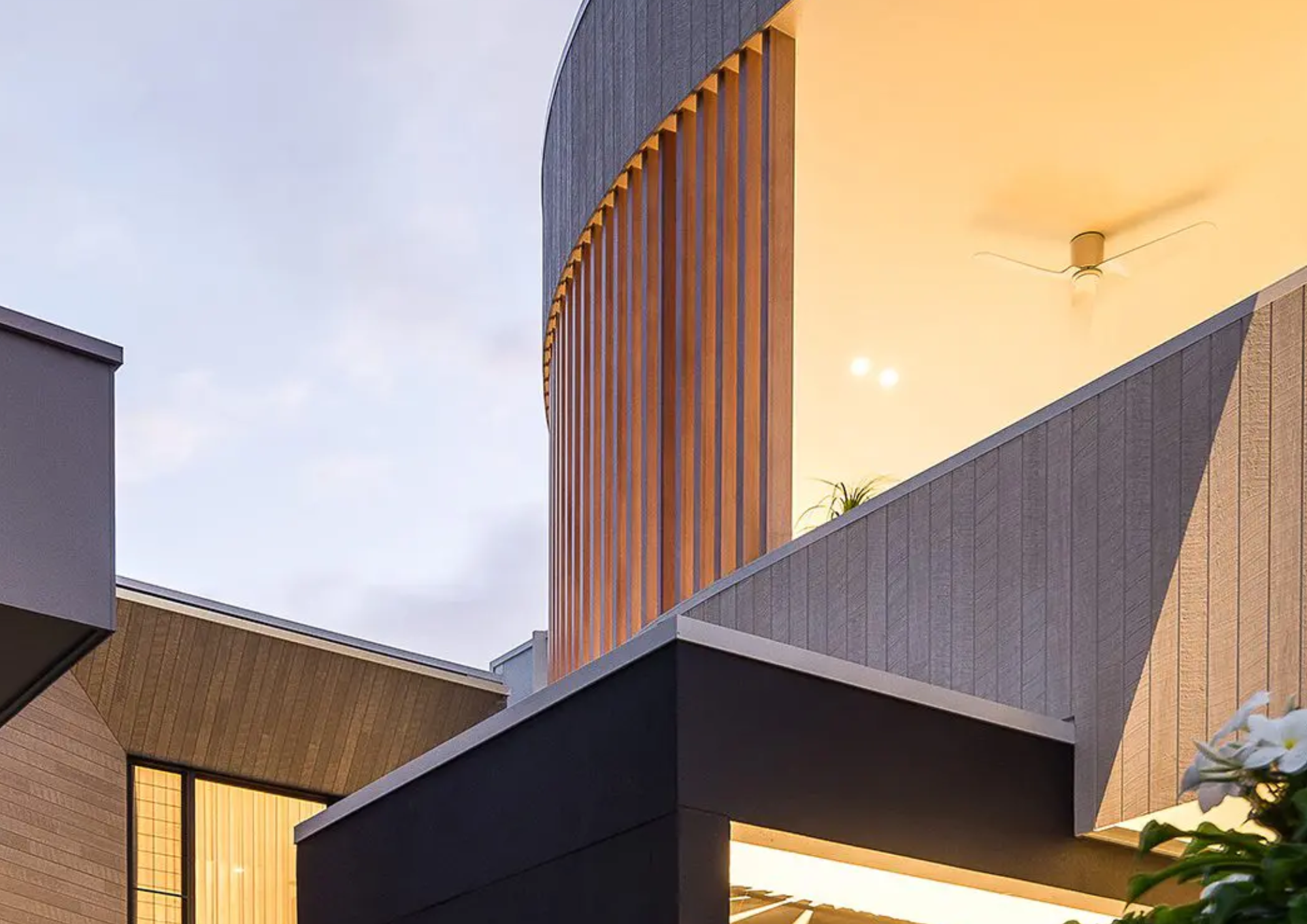 Modern building with angular design, featuring wood and gray panels, illuminated from within. The sky is clear with a few clouds visible.