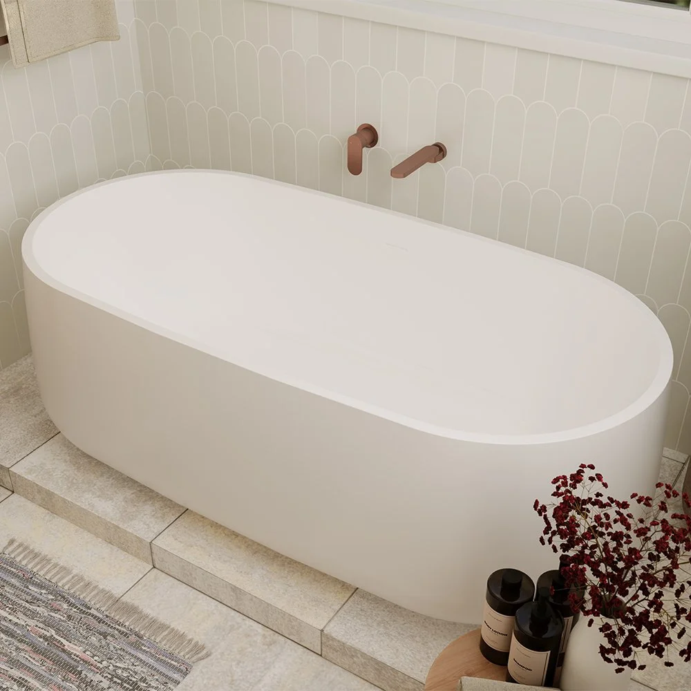 A modern, oval, white freestanding bathtub placed in a bathroom with light-colored tiled flooring and a patterned wall. There are copper-colored faucet fixtures on the wall above the bathtub, and some bottles and a decorative plant are on a small woo